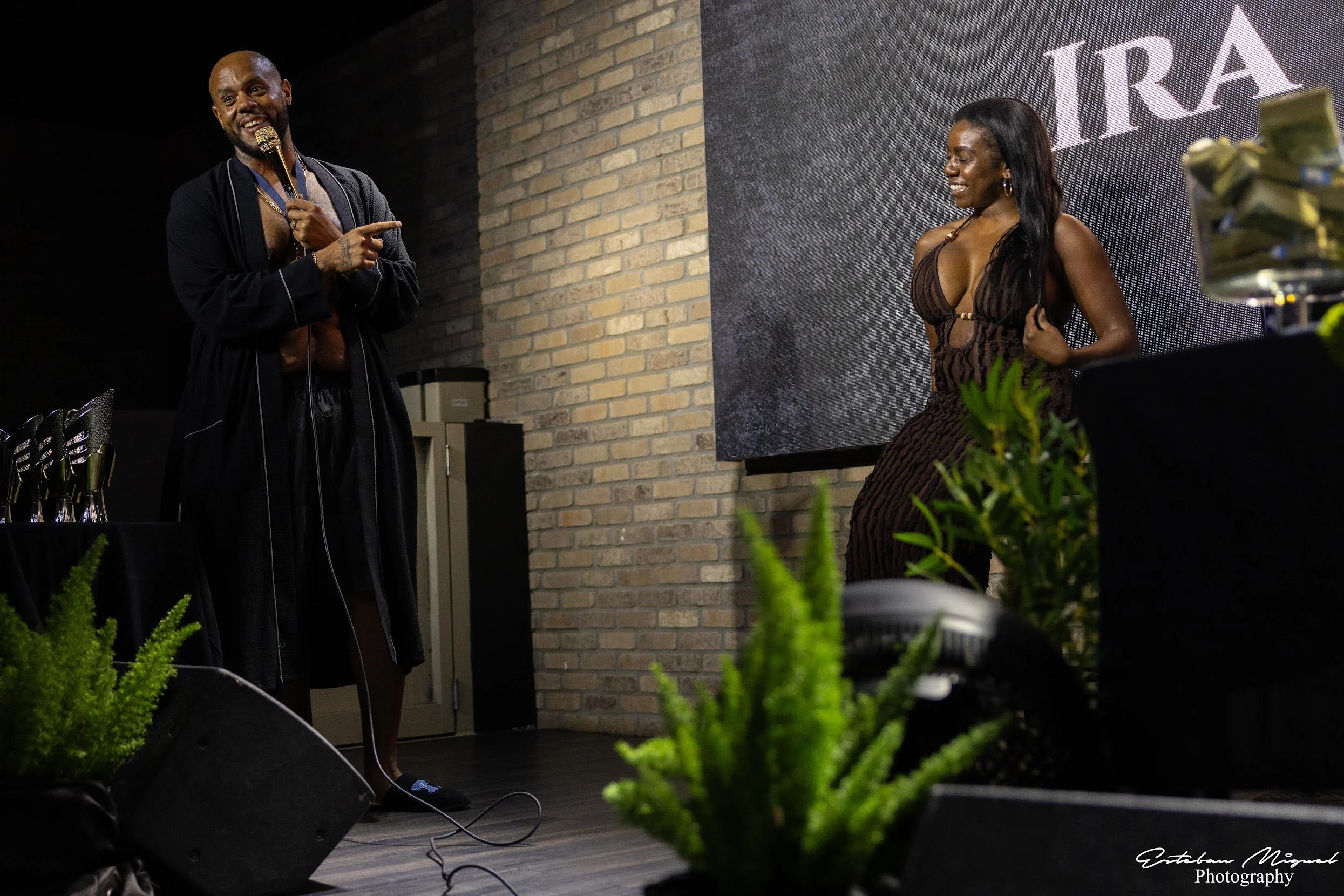 A man and a woman on stage at an event, with the man speaking into a microphone and the woman smiling. The background has a brick wall and a large screen displaying part of the word 'IRA'. There are awards or trophies on a table to the left, and some