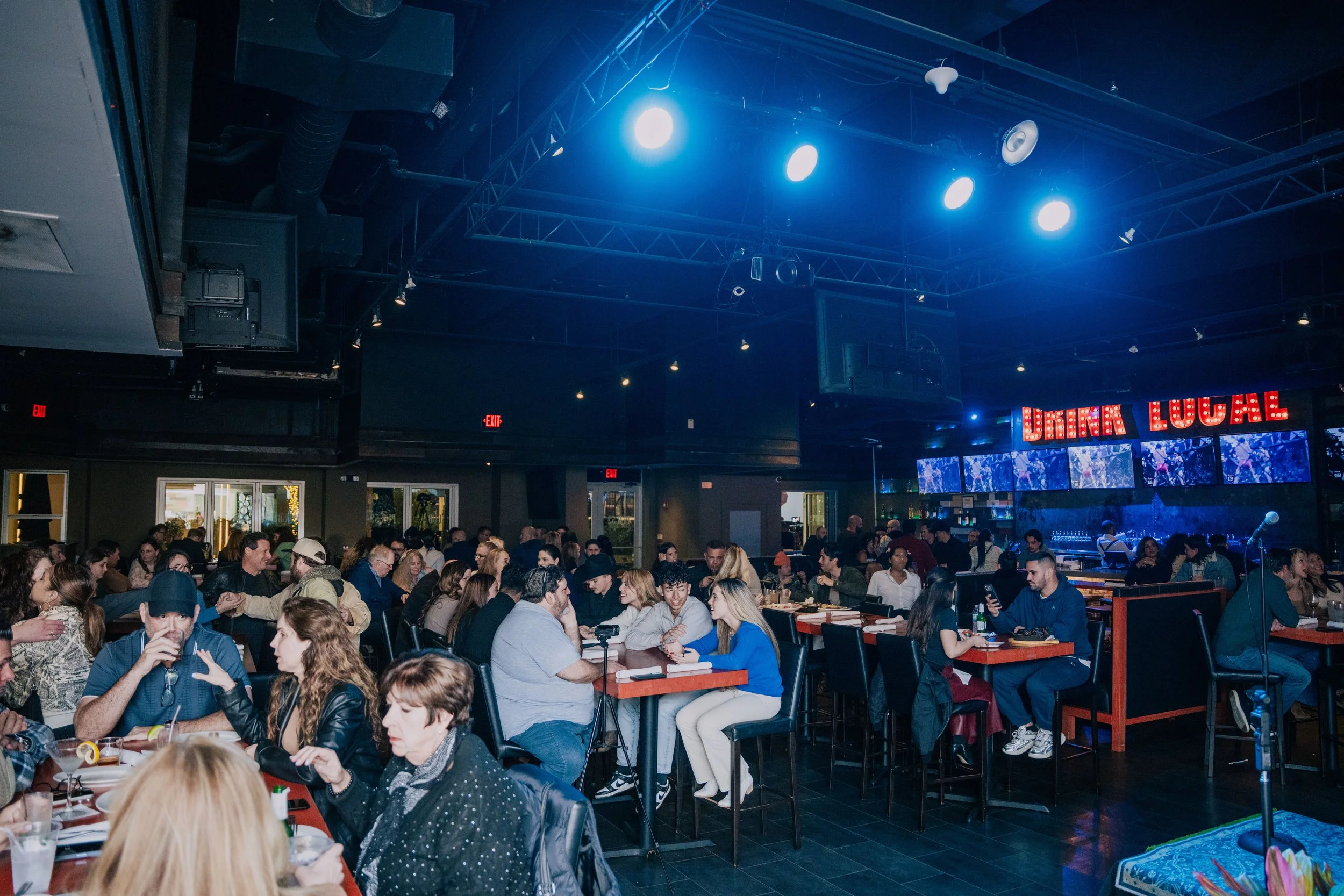 A crowded indoor venue with neon sign reading 'DRINK LOCAL', stage with blue lighting, and many people sitting at tables, socializing, and enjoying drinks.