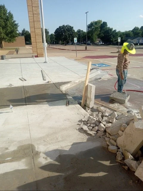 A construction worker wearing a yellow hard hat and camouflage shirt standing on a single concrete block at a construction site, with a pile of rubble and broken concrete in front of him. The background shows an empty parking lot and trees.