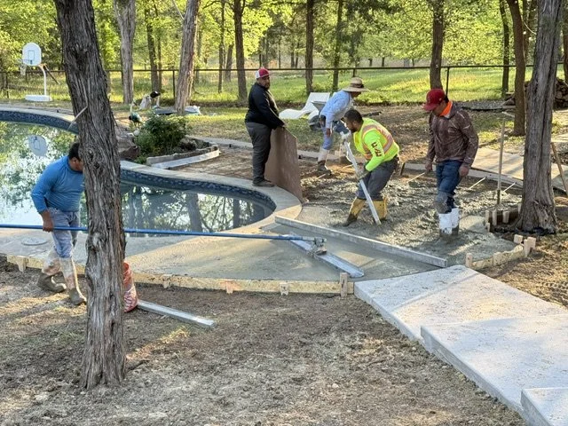 Construction workers are working on a mini-golf course, installing a new concrete pathway and surrounding the water hazard pool. They are using shovels and tools, and are surrounded by trees and a fence.