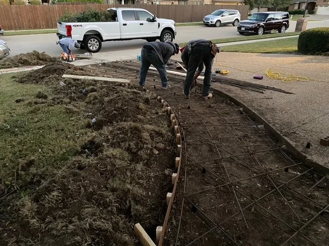Two workers are installing a curved concrete curb on a sidewalk with rebar reinforcement, surrounded by dirt and construction tools, near parked cars on a residential street.