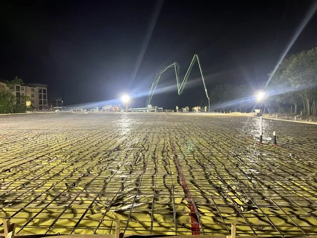 Nighttime construction site with bright lights, steel rebar, and yellow insulating material, with a large illuminated crane or lifting structure in the background.