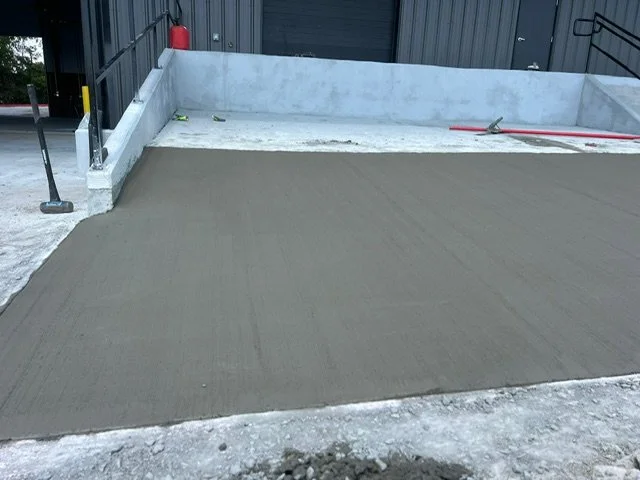 Newly poured concrete slab in a parking area, with a concrete barrier and construction tools nearby.