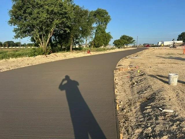Freshly paved asphalt road under clear blue sky with trees and construction materials on the side.