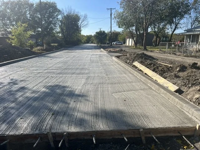 Newly paved road under construction with curb work in progress on a sunny day, trees with changing leaves in the background.