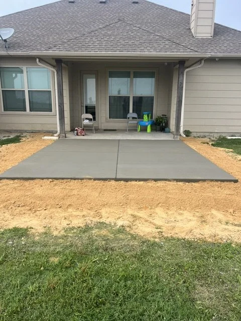 Backyard patio with newly poured concrete slab and surrounding dirt, outdoor chairs, and children's toys near the house.