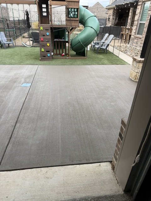 View of a backyard play area with a playset, green slide, and climbing wall, as seen from inside a house through a sliding door, with concrete patio and neighboring houses in the background.