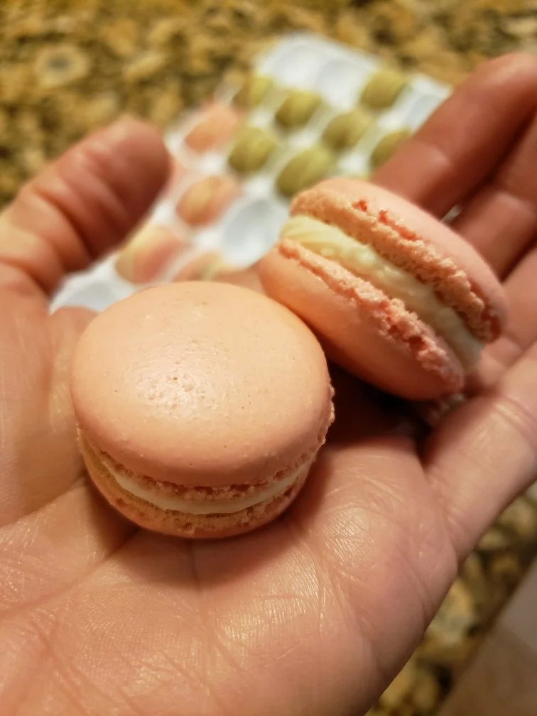 Close-up of a person's hand holding two pink macarons with a creamy filling, with a tray of similar macarons in the background on a granite countertop.