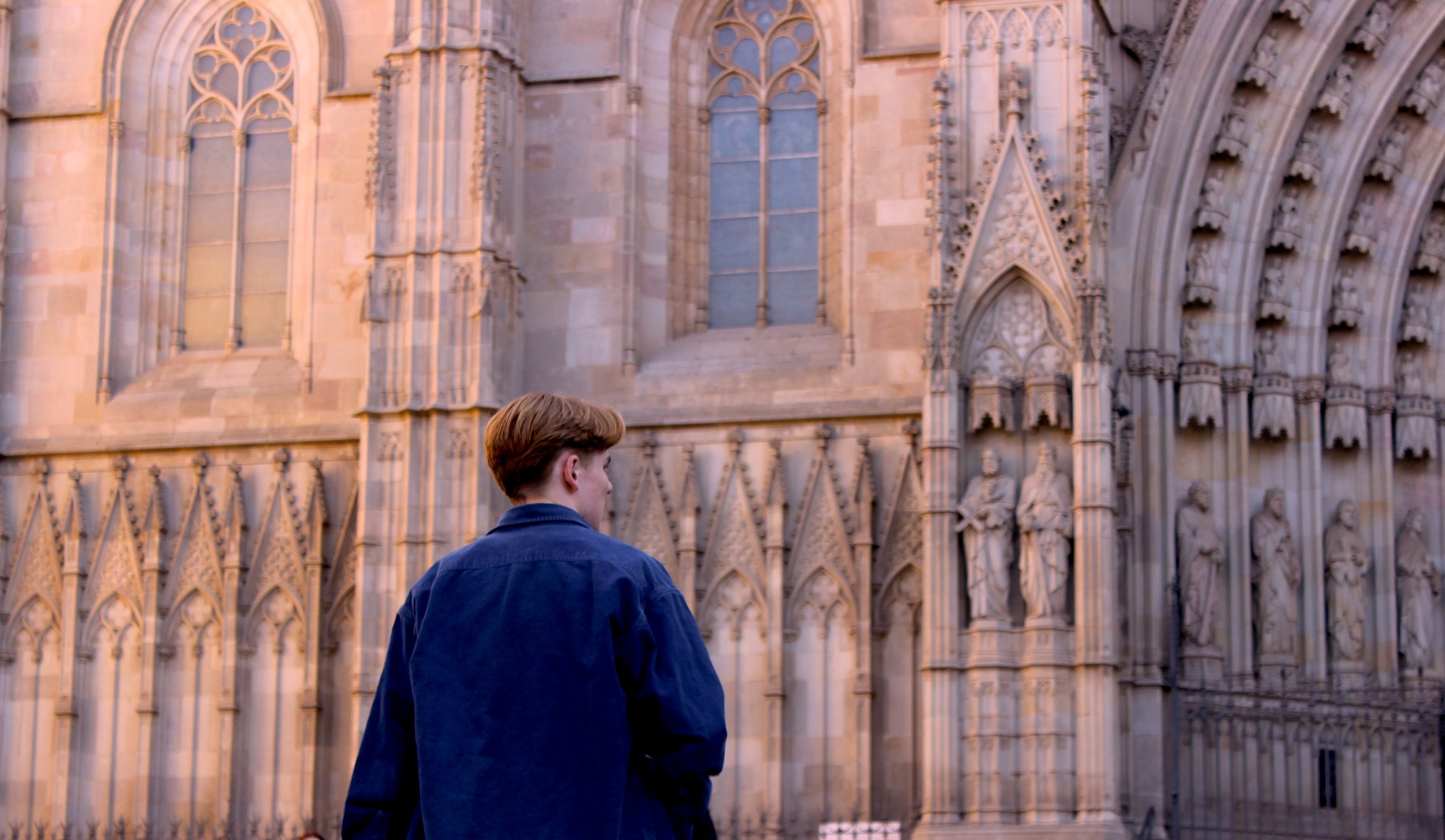 A person with light brown hair wearing a dark blue jacket standing in front of an ornate stone gothic cathedral with tall arched stained glass windows and detailed sculptures on the facade.