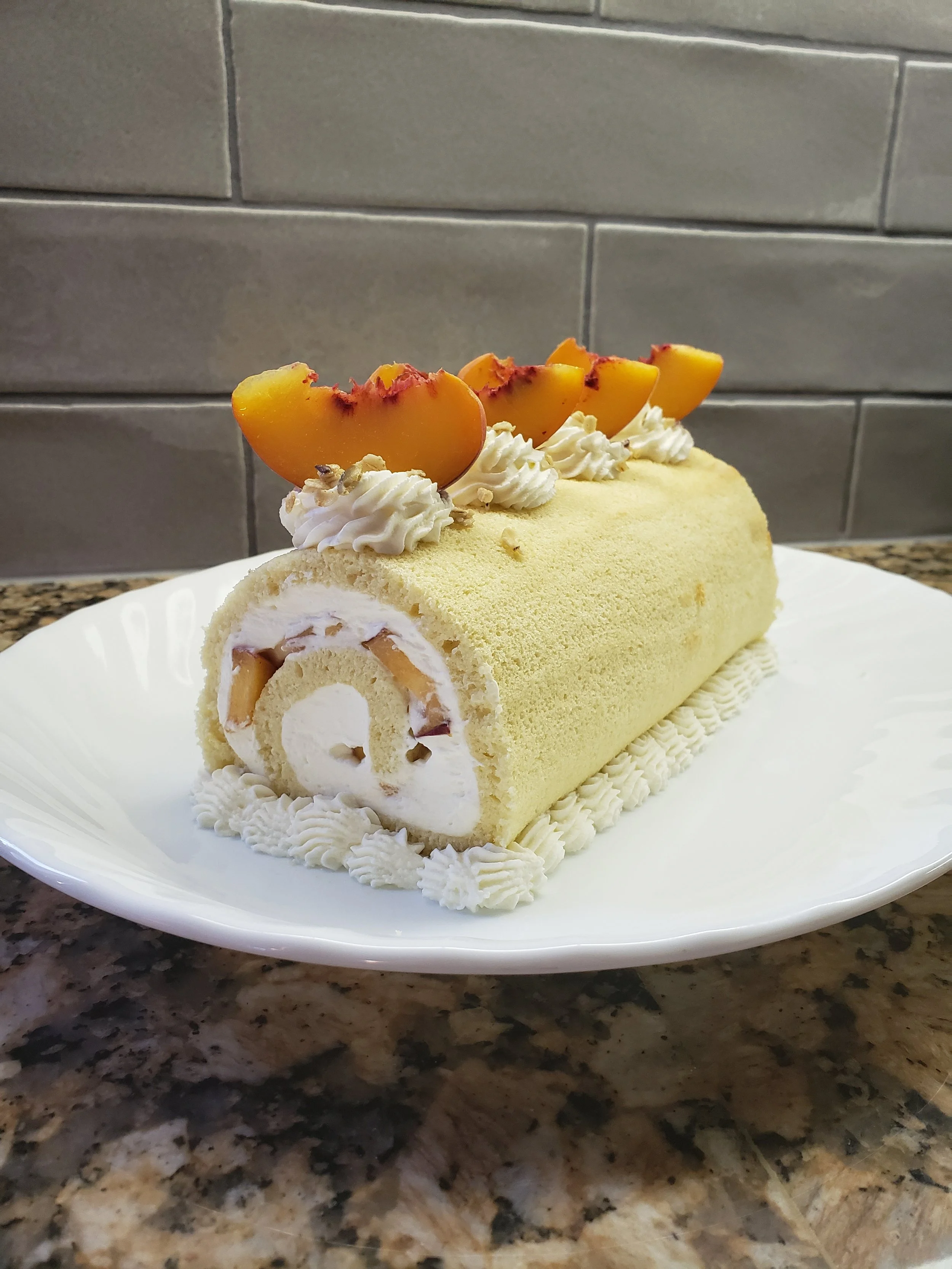 A Swiss roll cake with yellow sponge, white cream filling, and peach slices on top, decorated with whipped cream and chopped nuts, served on a white plate.