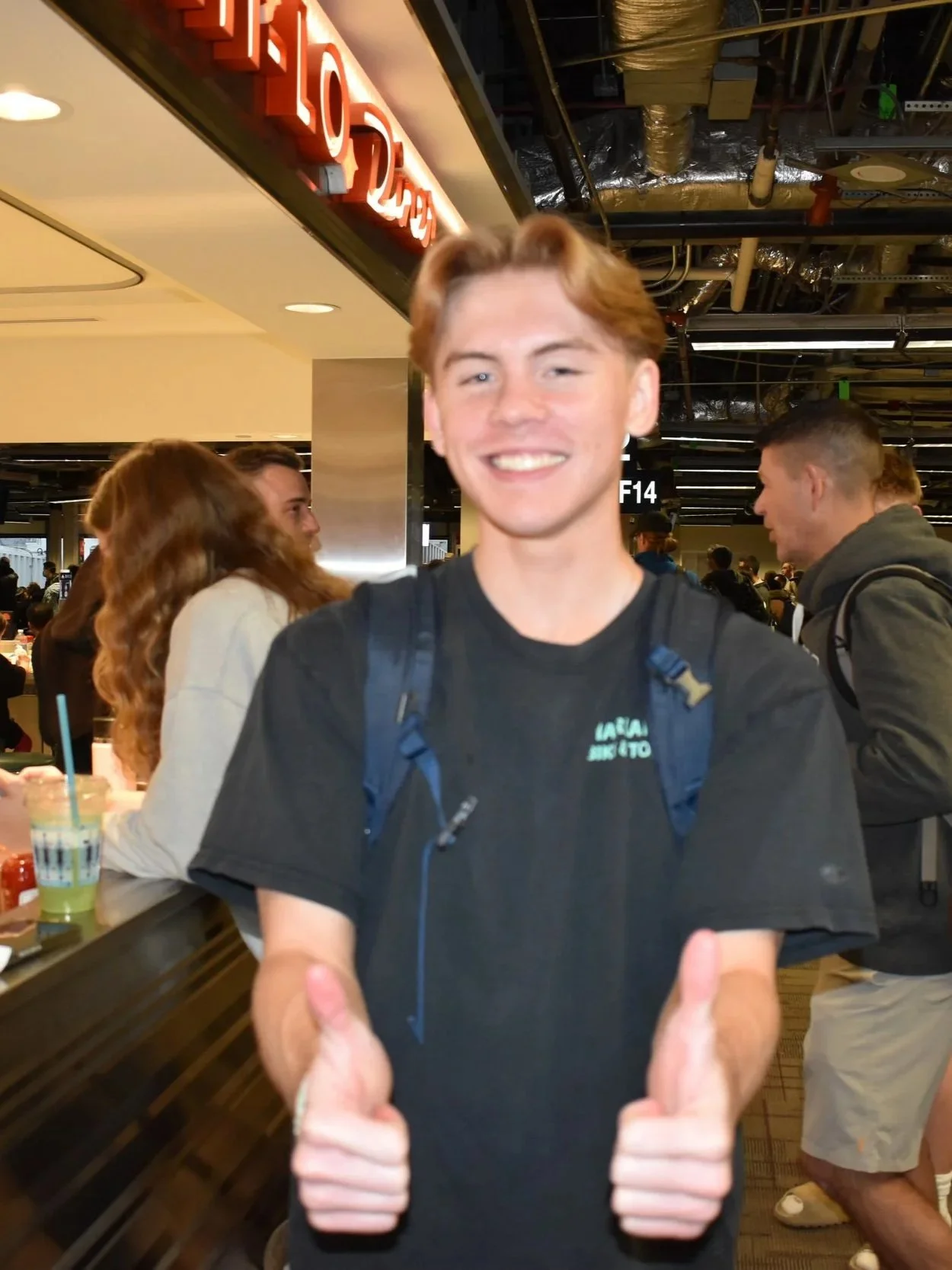 A young man with red hair smiling at the camera while giving two thumbs up in a busy indoor setting, likely an airport or shopping mall, with people in the background and visible ceiling pipes and lights.