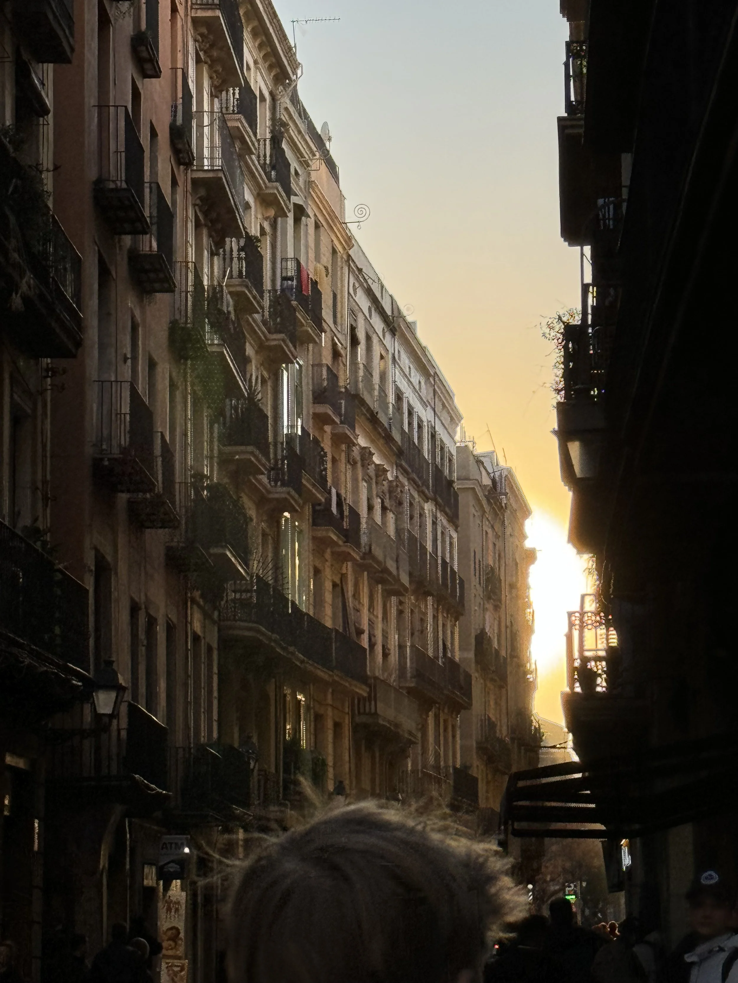 A street scene during sunset with 4 to 5 story European-style buildings on both sides, featuring balconies, some with plants, and streetlights. There are many people walking in the street, and the sun is setting in the background, casting a warm glow over the scene.