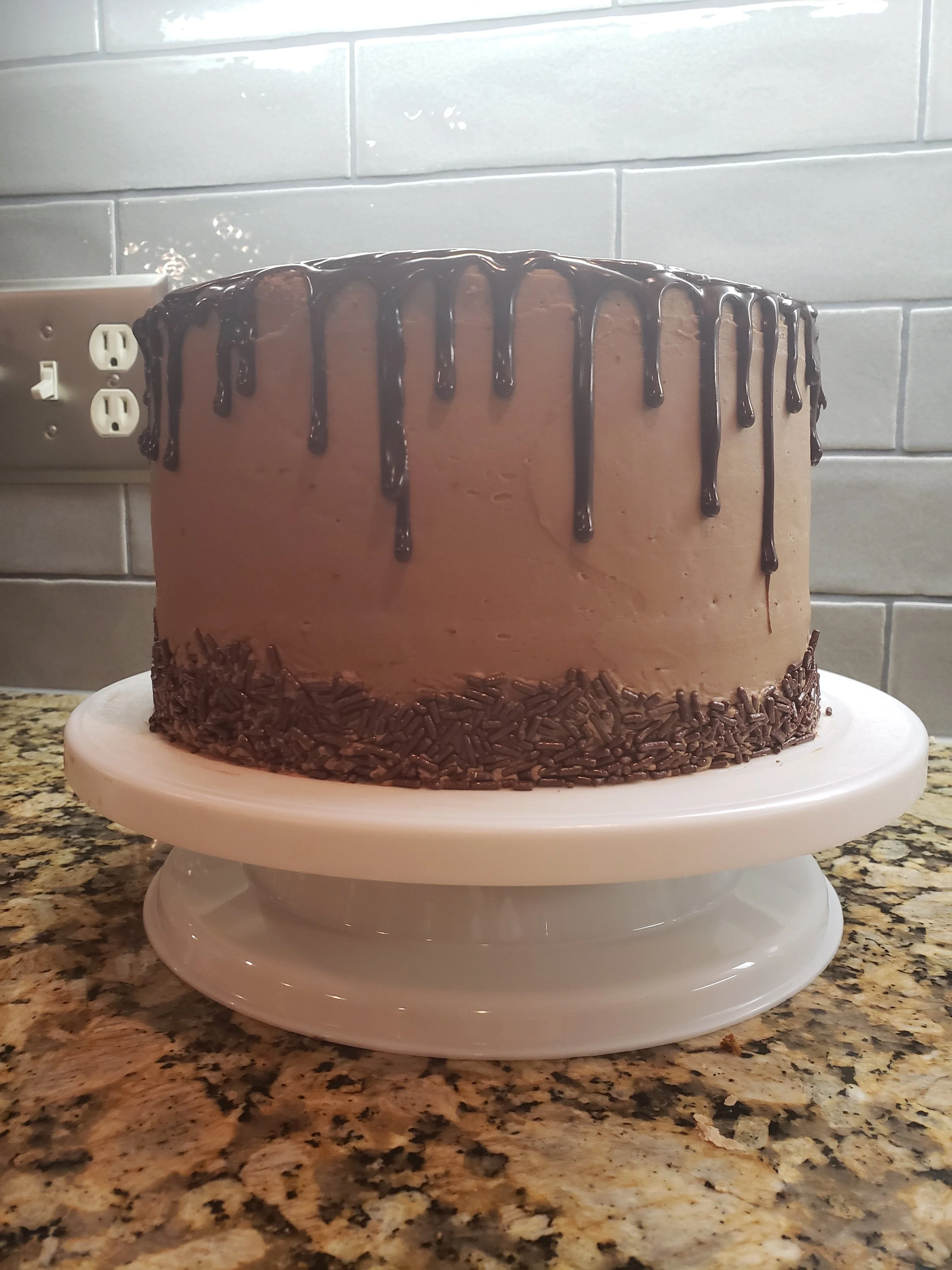 A chocolate frosted cake with chocolate drizzle on top, placed on a white cake stand on a kitchen countertop with a grey tile backsplash and an electrical outlet.