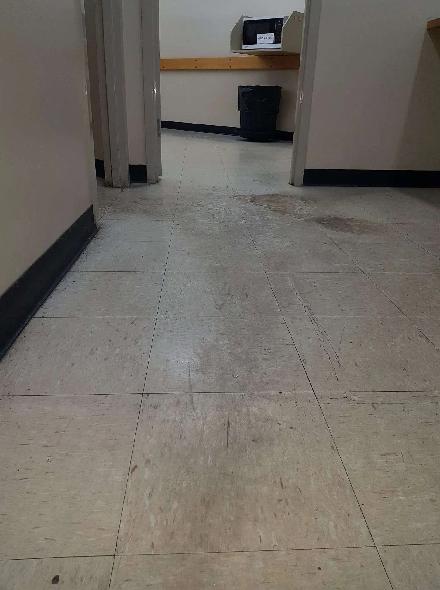 Clean, light-colored tiled floor with some dirt and scratches, leading to a doorway. Inside the room, there is a black trash bin, a wooden shelf, and a microwave on top of the shelf. The walls are plain, and part of a shelf is visible.