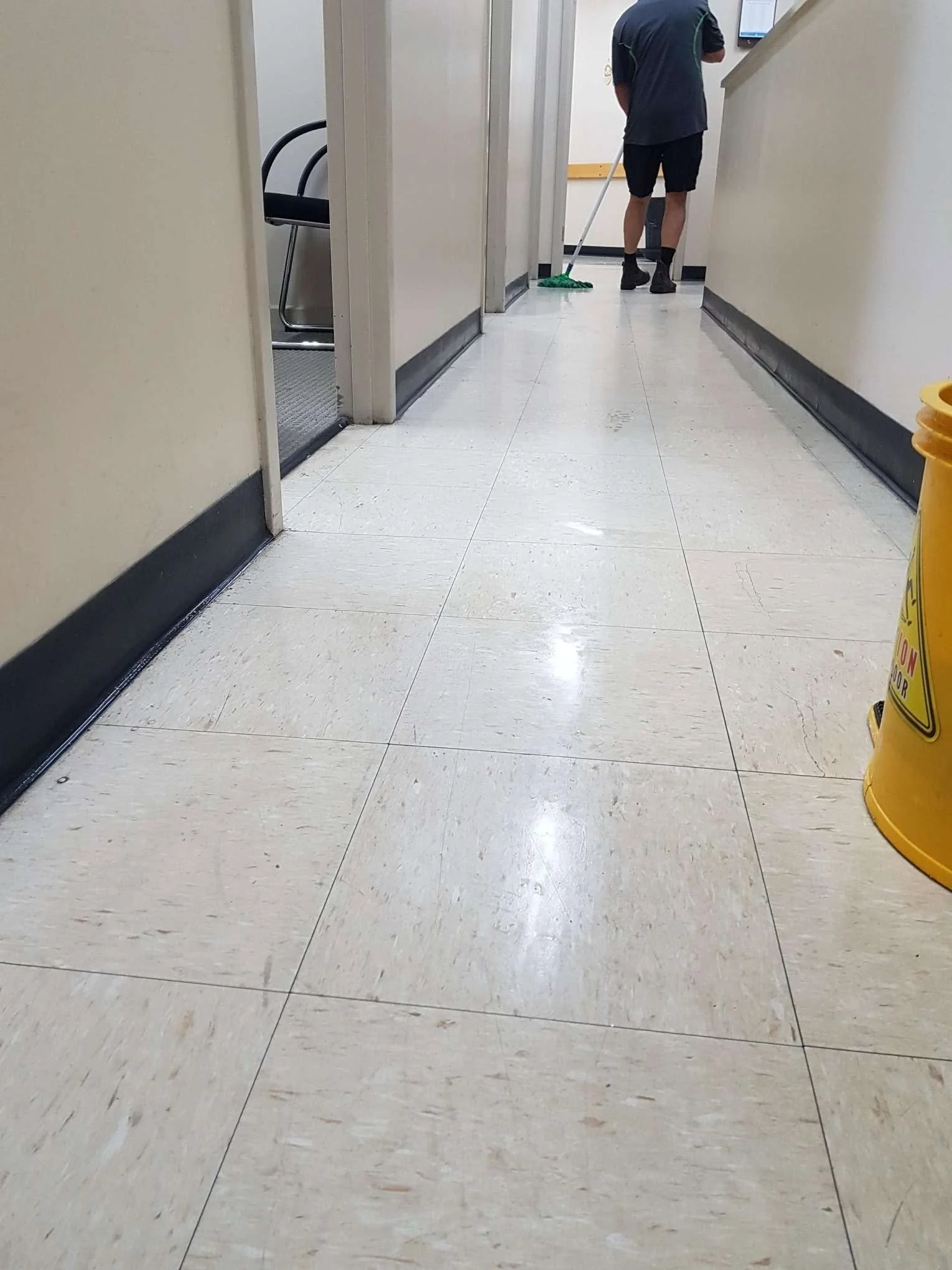 A person mopping the floor in a hallway, wearing dark shorts, a dark shirt, and black boots, with a monitor on the wall in front of them and a chair visible through an open door on the left.