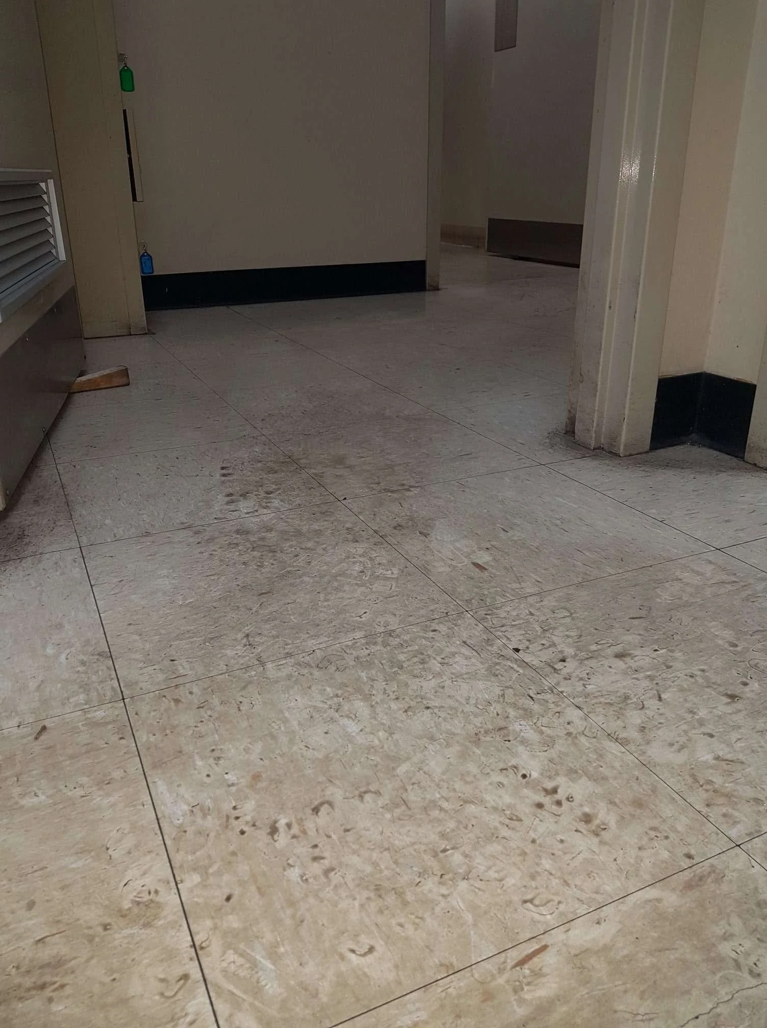 Dirty beige tiled floor with smudges, stains, and debris. Part of a wall and a door are visible in the background.