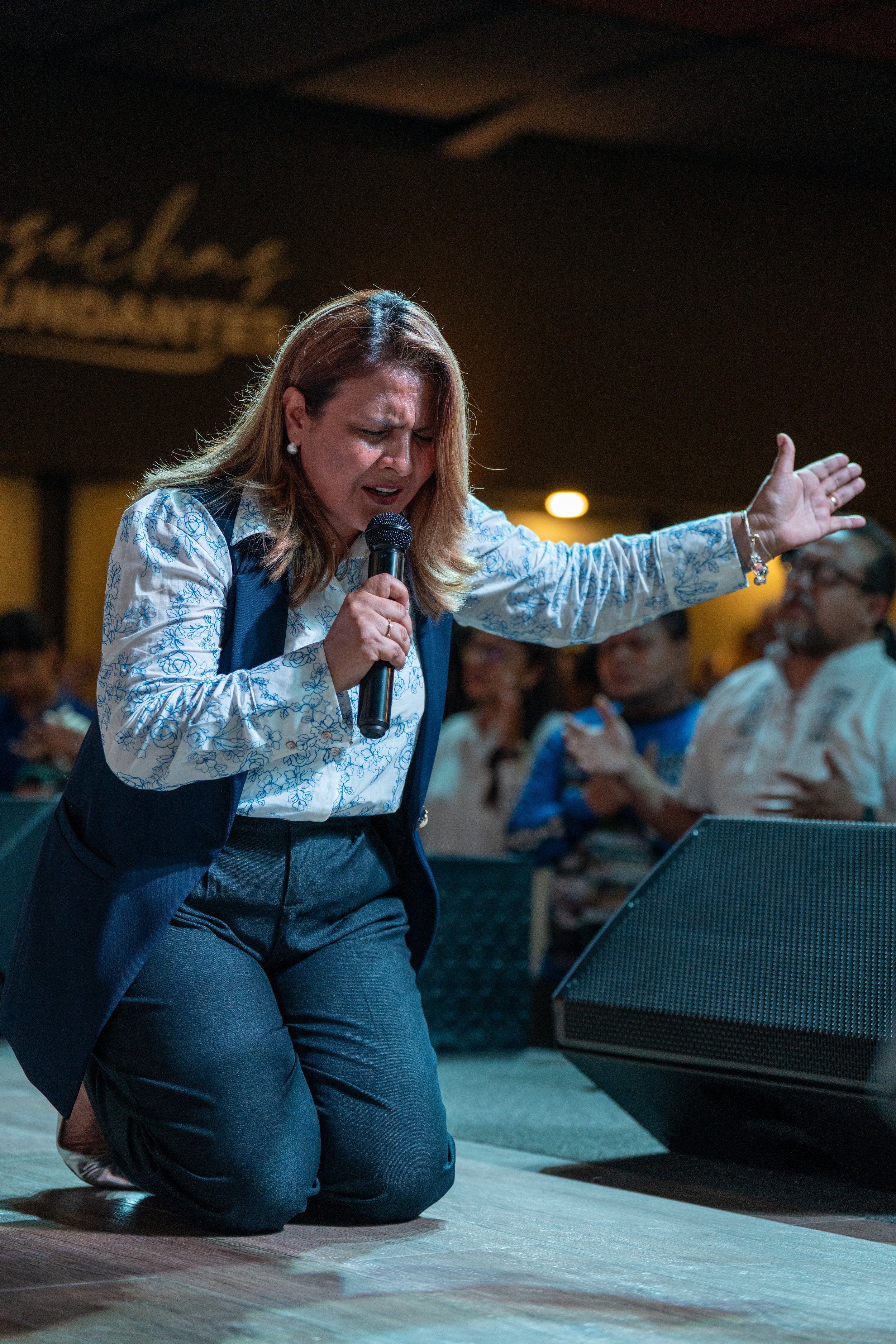 Mujer orando con los ojos cerrados y una mano extendida frente a un micrófono en un evento congregacional.