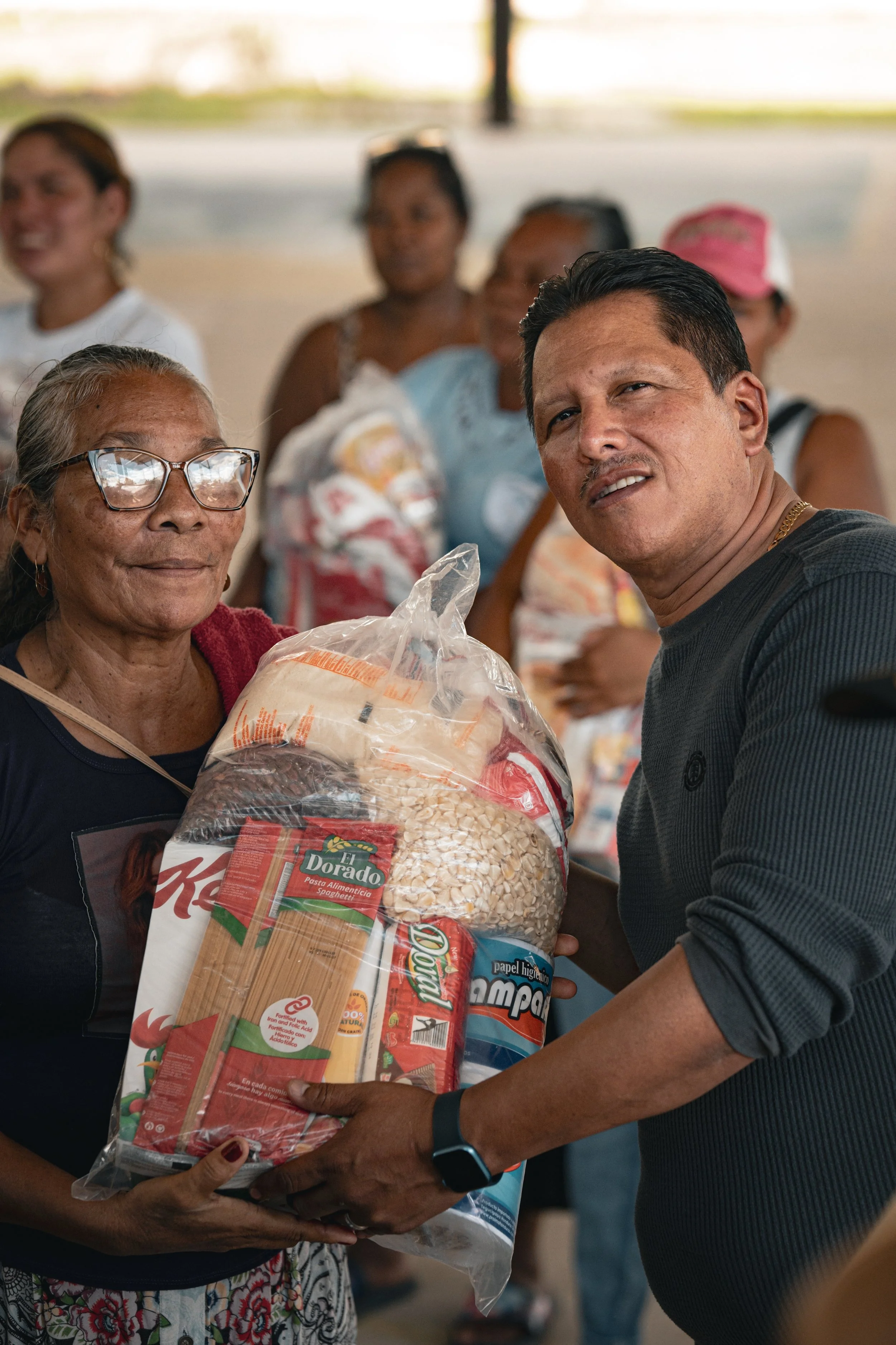 Una mujer de edad avanzada recibe un paquete de alimentos de un hombre, en un entorno que parece ser un evento comunitario o mercado.