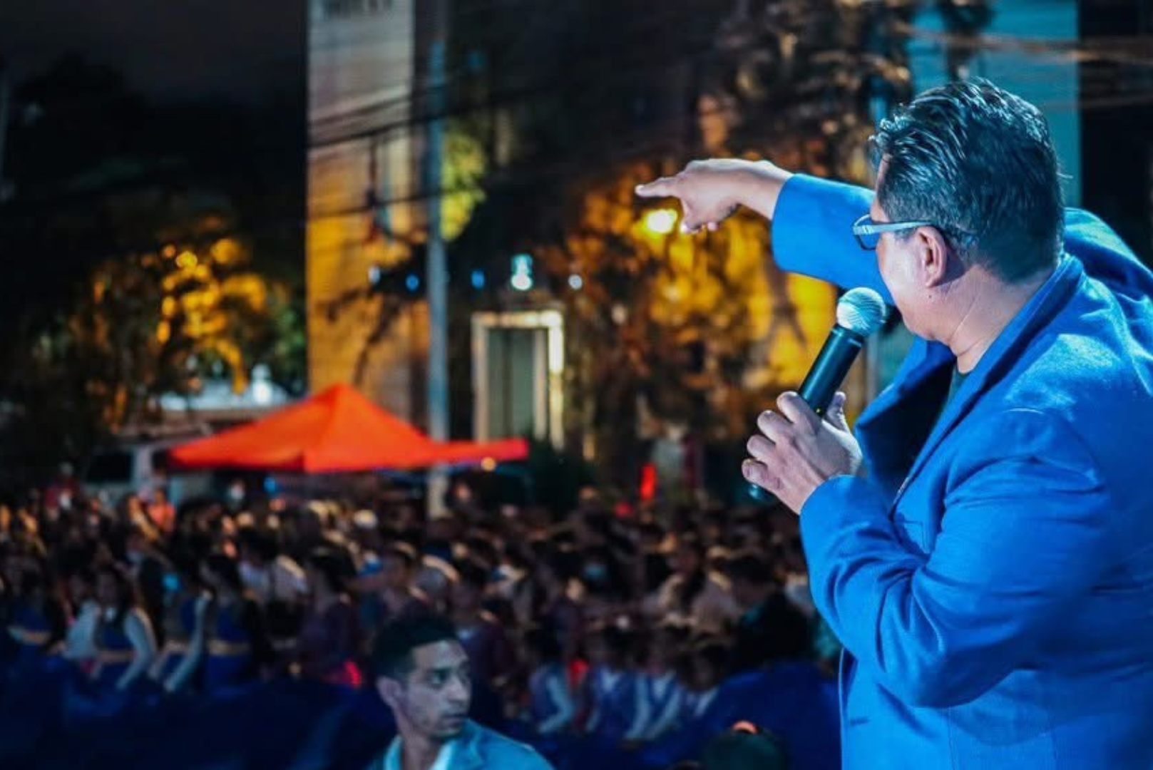 Hombre con gafas y chaqueta azul hablando en un micrófono, señalando con su mano hacia una multitud en una calle iluminada durante la noche
