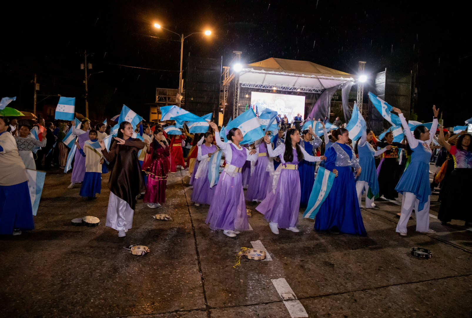 Grupo de personas participando en un evento nocturno, con mujeres vestidas con trajes largos en tonos morados, azules y blancos, bailando y sosteniendo banderas de Honduras. En el fondo, hay una tarima con un escenario iluminado y un grupo de músicos
