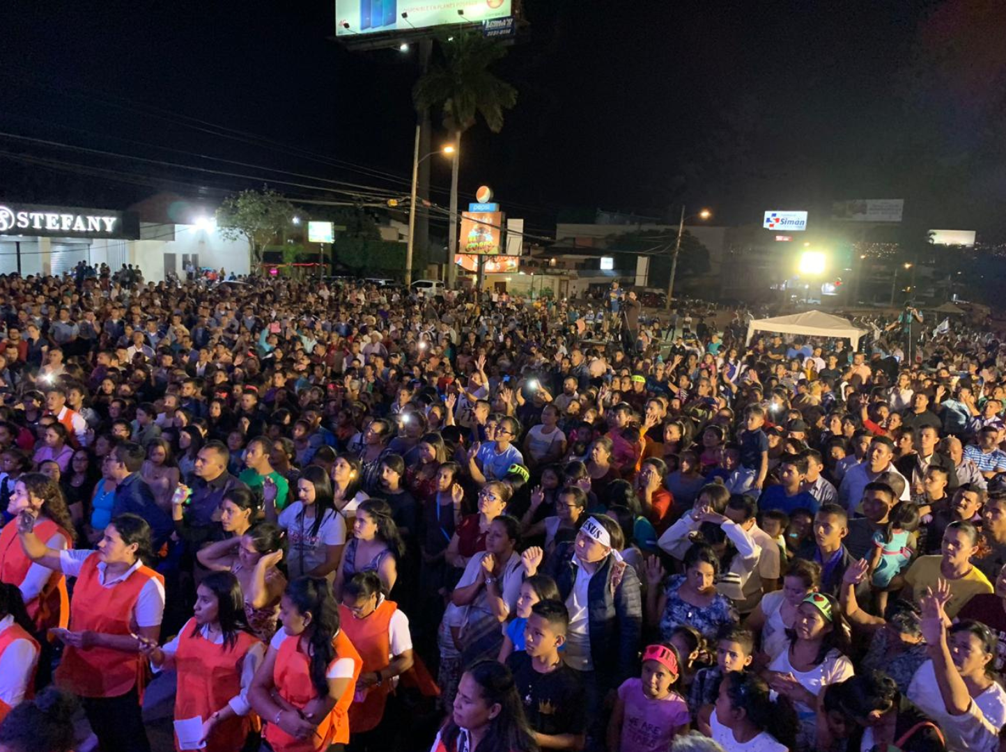 Una multitud de personas reunidas en un evento nocturno, muchas de ellas levantando las manos, algunos con camisetas o uniformes, en un espacio abierto con cartelera y anuncios luminosos de fondo.