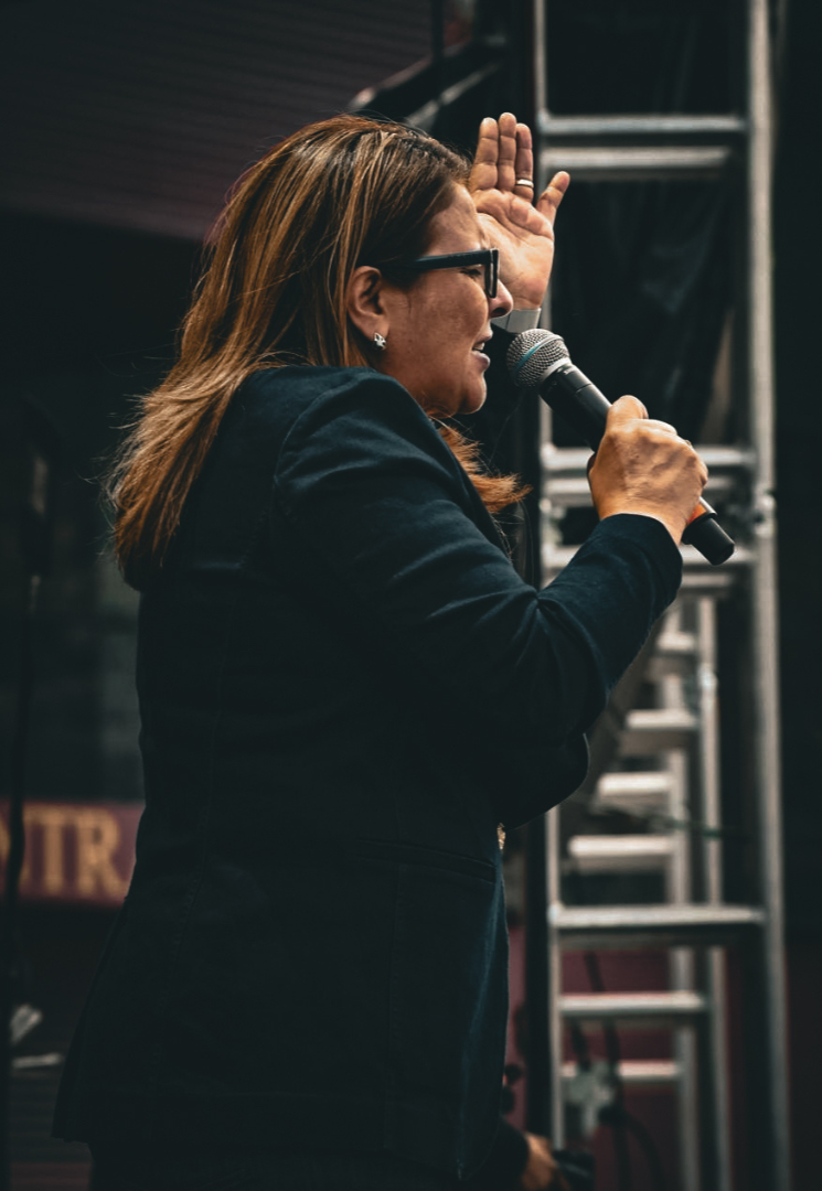 Una mujer con gafas y cabello castaño, vestida con saco negro, hablando a un micrófono en un escenario, levantando su mano en señal de énfasis.
