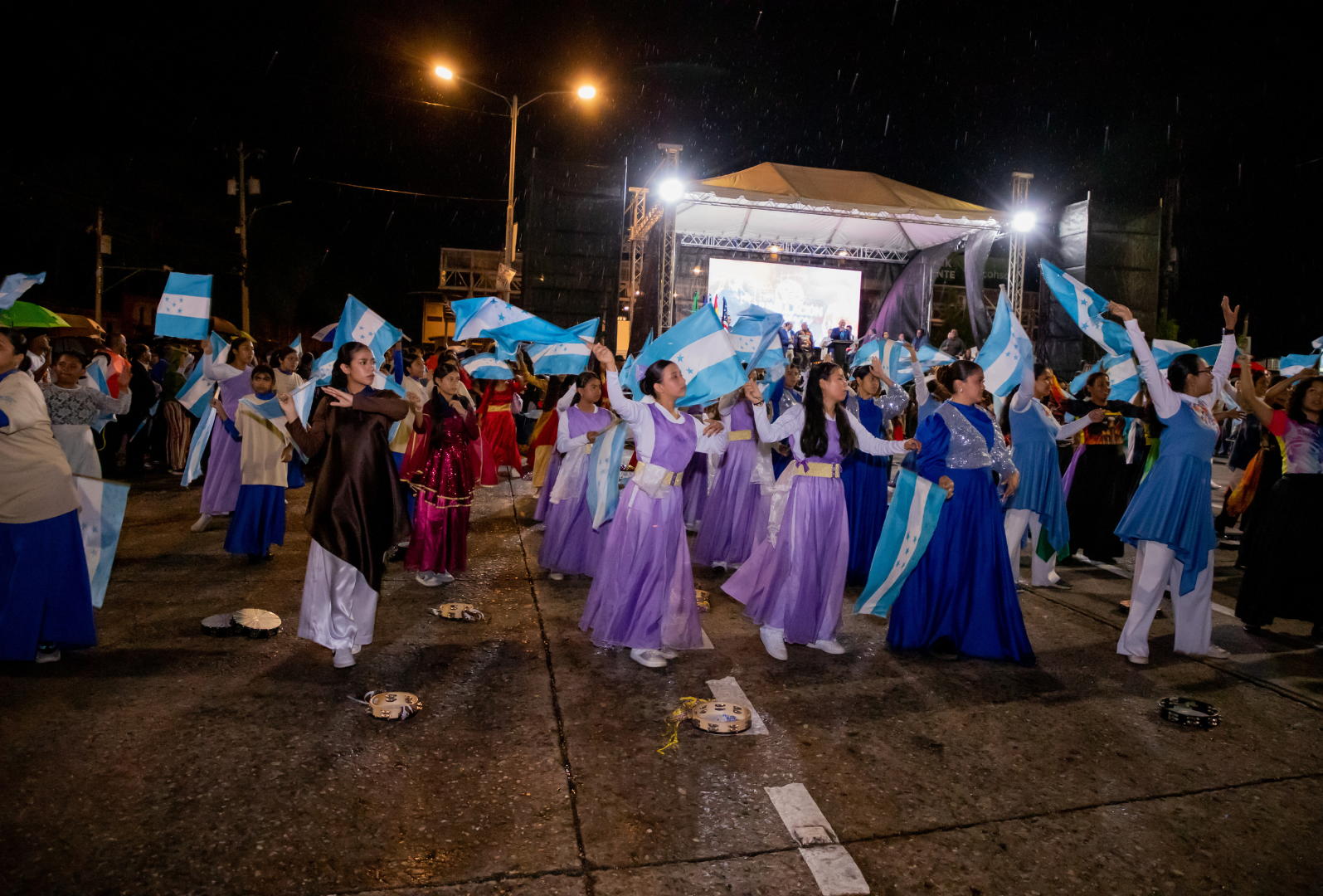 Celebración en la calle con baile de personas vistiendo trajes tradicionales grandes banderas de Honduras, en una noche lluviosa con escenario y luces en el fondo.