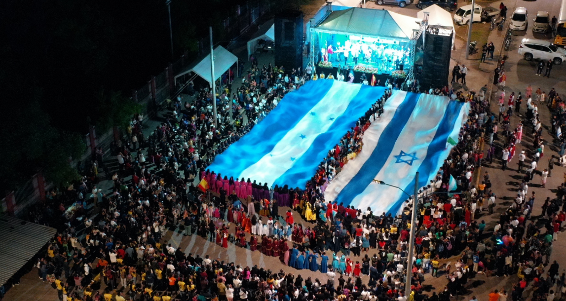 Celebración pública con una gran bandera de Argentina y otra de Israel en una plaza rodeada de muchas personas.