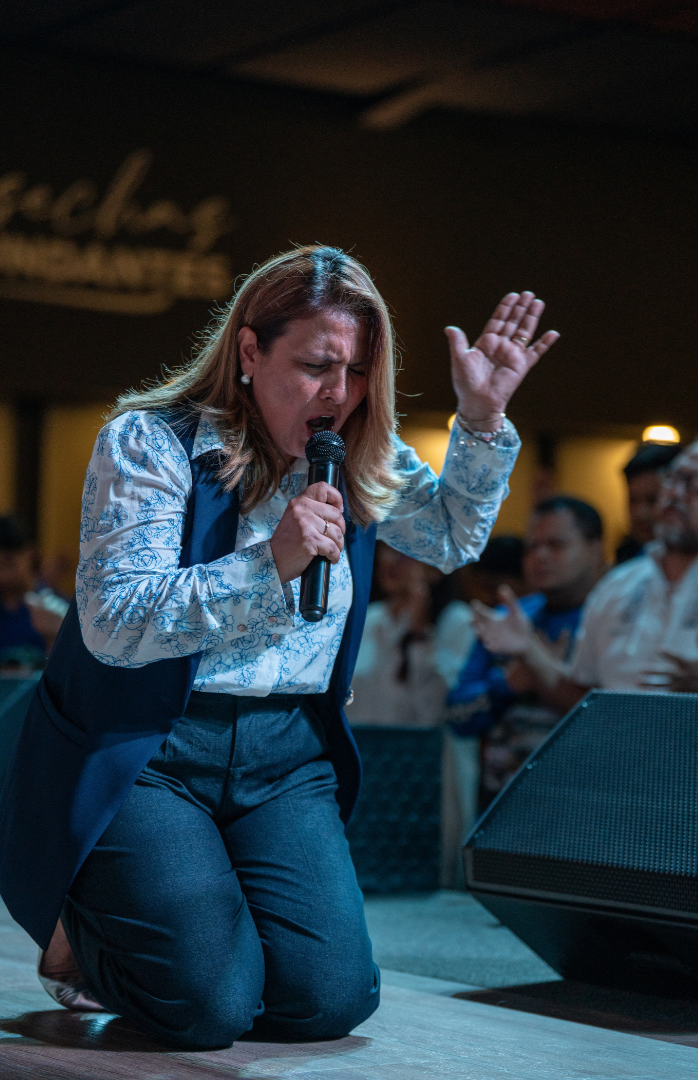 Mujer orando con un micrófono en la mano, arrodillada con una expresión de devoción en un entorno de iglesia o centro de culto.