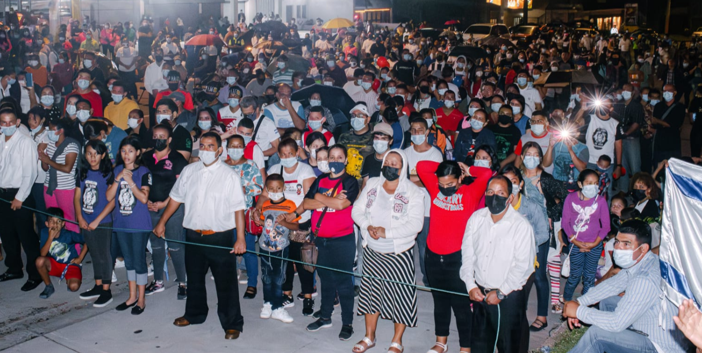 Gran multitud de personas en una protesta o manifestación al aire libre durante la noche, muchas con mascarillas y algunos con paraguas, en una calle urbana.
