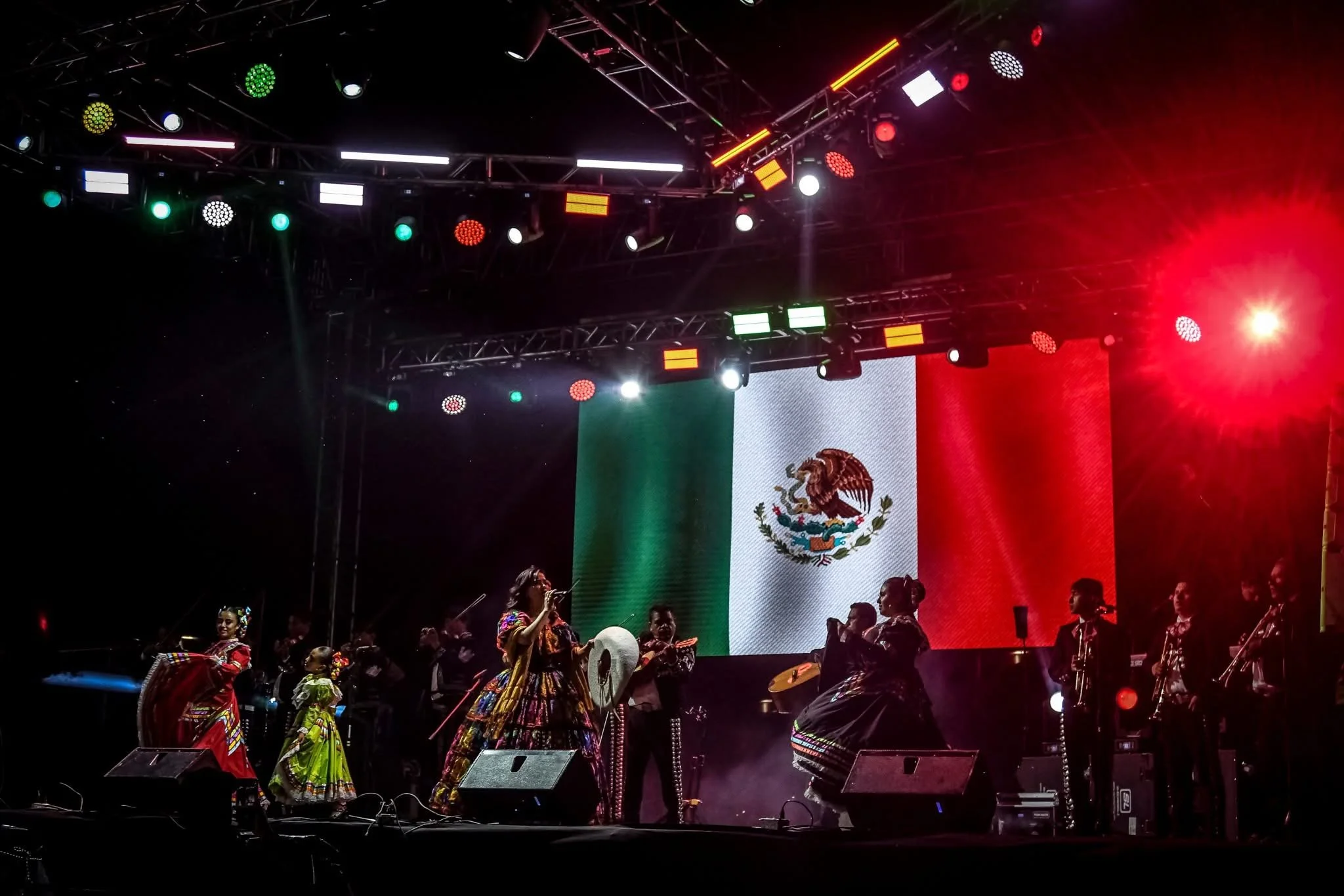 Espectáculo en un escenario con banderas mexicanas y grupos de personas con vestimenta tradicional mexicana, mientras hay luces de colores y una pantalla gigante de fondo.