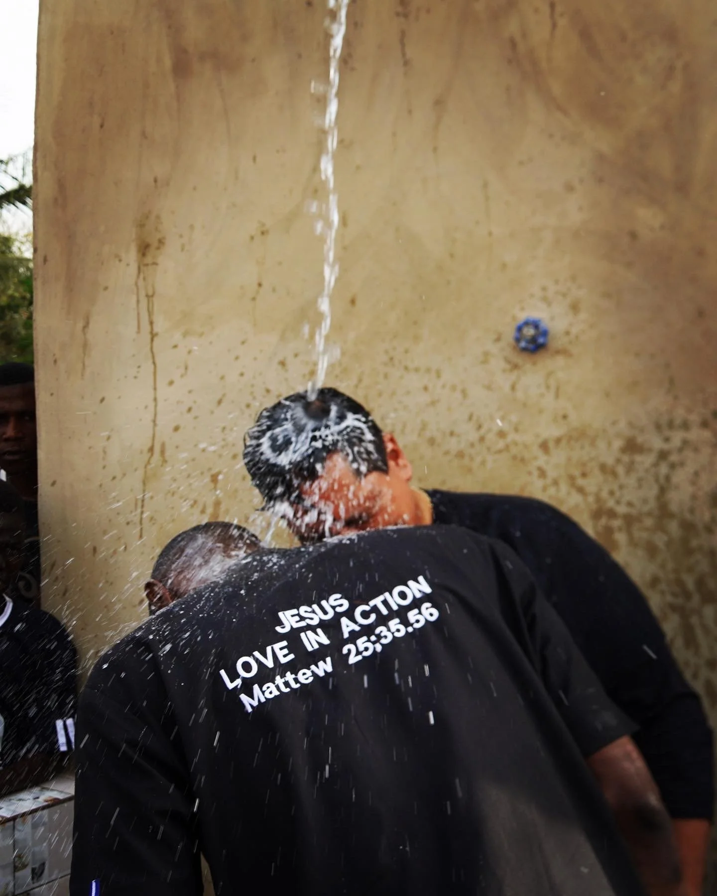 Un grupo de personas en un acto religioso, una de ellas con una camiseta que dice 'JESÚS, LOVE IN ACTION, Matthew 25:35-36', en un lugar con pared de fondo beige y una teja en la pared.