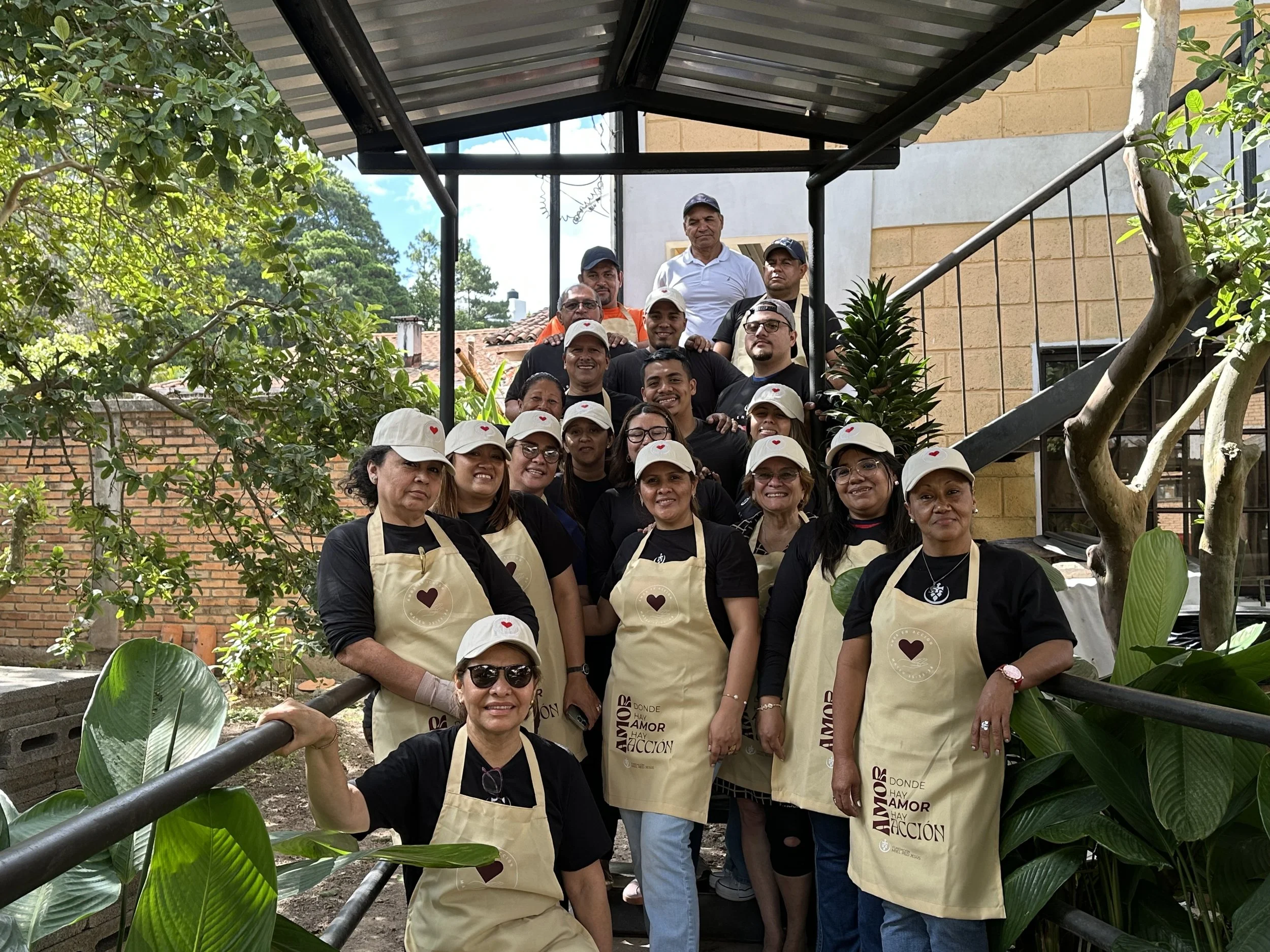 Grupo de personas jóvenes y adultas posando en un espacio al aire libre, algunas llevan delantales beige con texto en español y gorras blancas con corazones rojos, rodeados de plantas y árboles, con una estructura de sombra en el fondo.