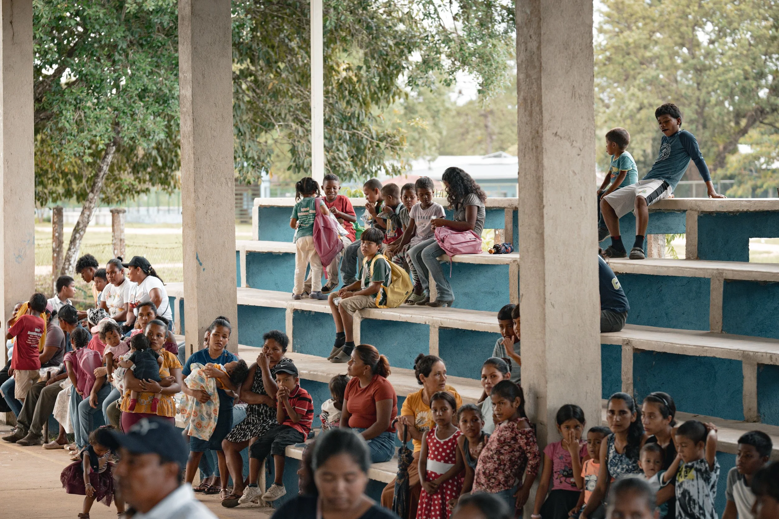Niños y adultos sentados en asientos de un espacio abierto con columnas, rodeados de árboles y naturaleza.