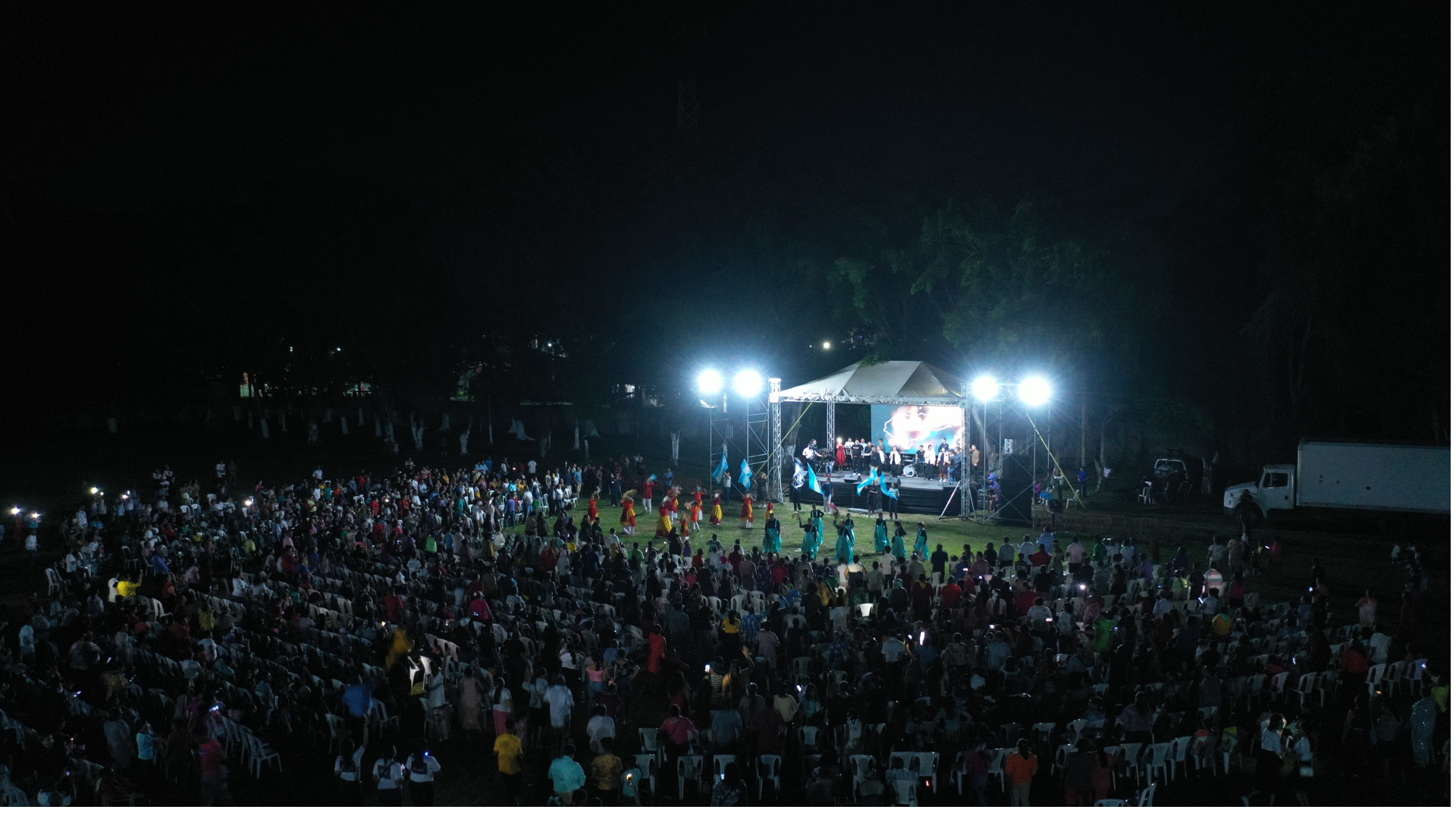 Concierto al aire libre por la noche con una multitud de personas, un escenario con músicos y artistas, iluminación intensa y un ambiente festivo.