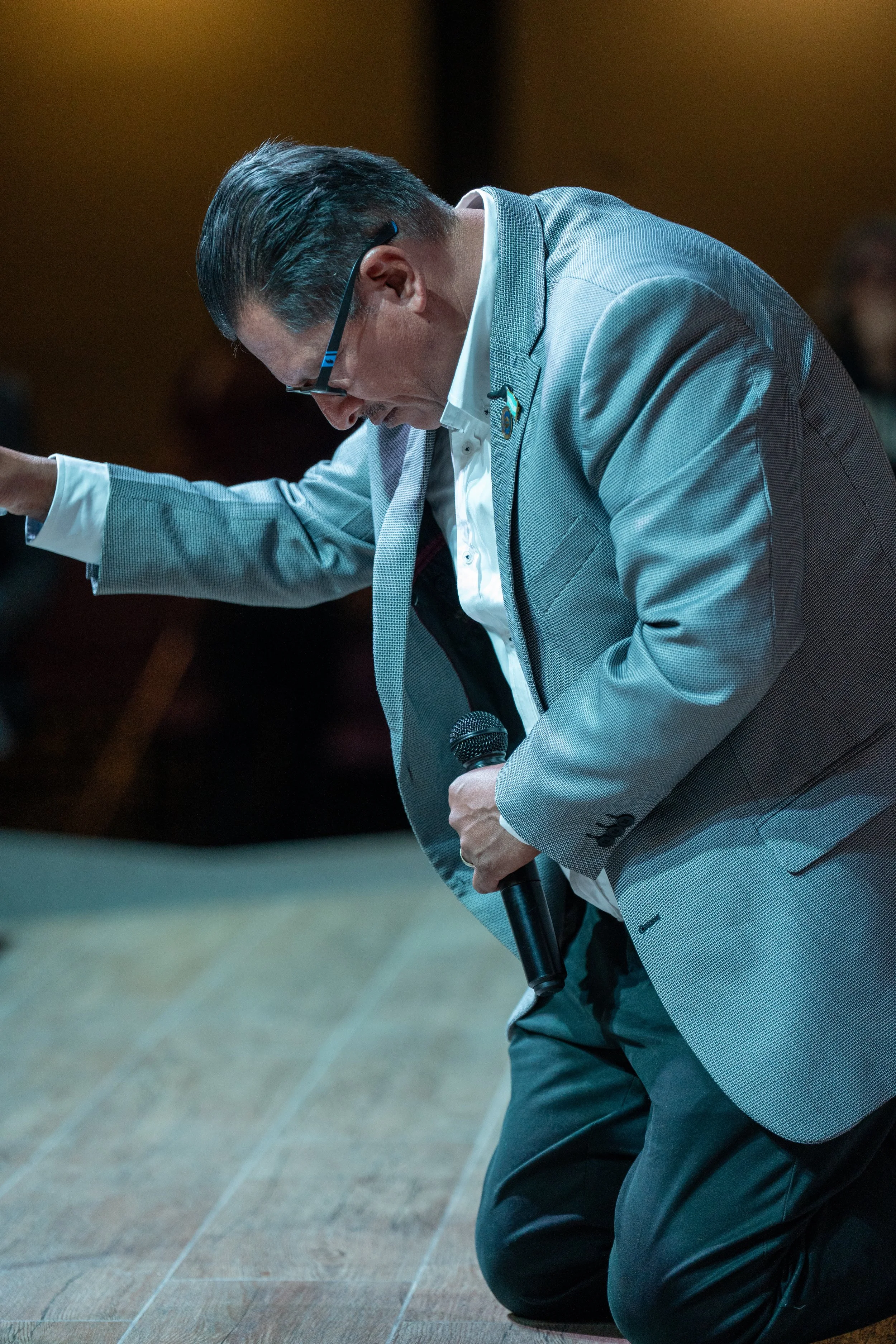 Hombre con gafas y traje arrodillado en oración en un lugar interior, en actitud de respeto o espiritualidad.