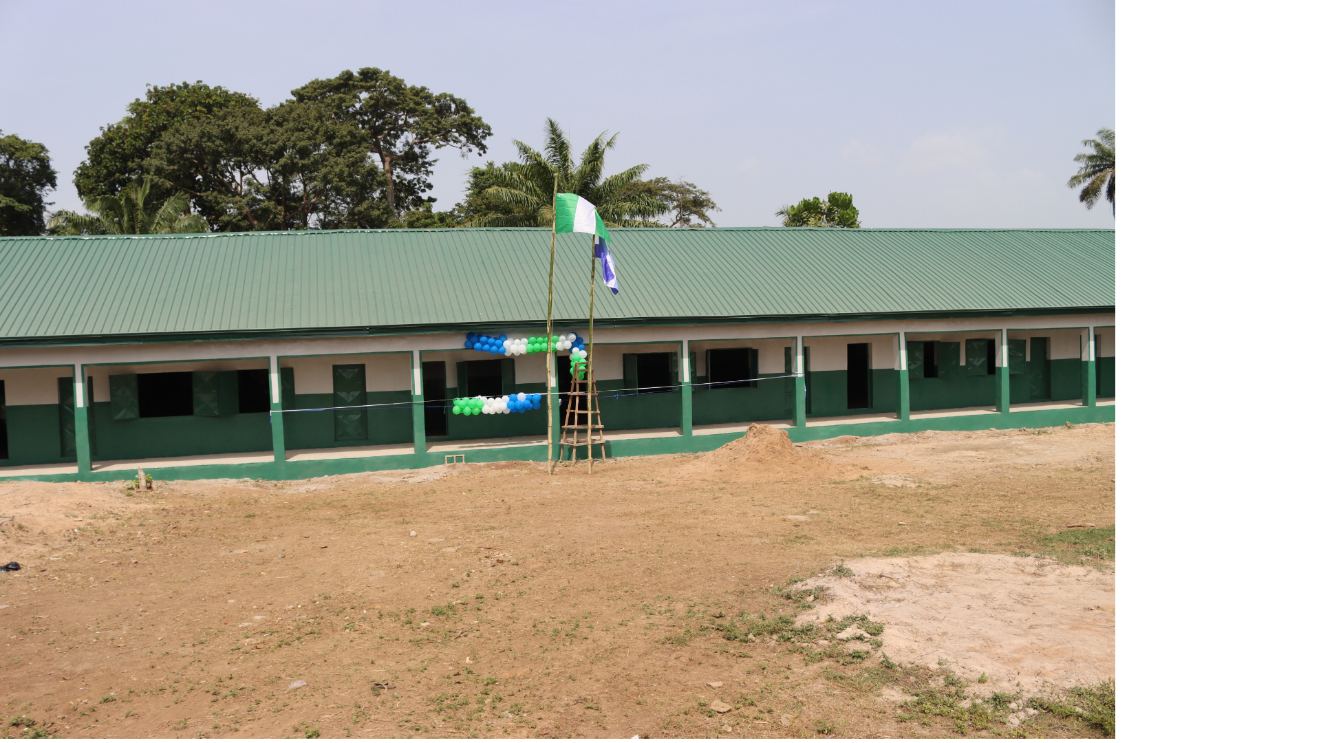Construcción de un edificio escolar con 12 aulas, banderas en una asta en el centro, adornada con globos en los colores nacionales, en un campo de tierra con árboles tropicales al fondo.