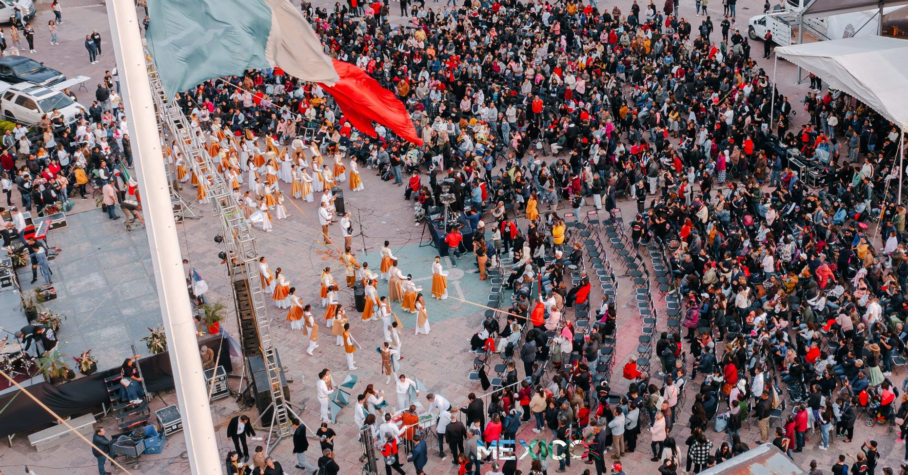 Gran multitud de personas en una plaza pública durante un evento cultural. Al centro, un grupo de bailarines en trajes tradicionales mexicanos realizando una presentación. Al fondo, una gran bandera de México ondeando, y a la derecha varias carpas blancas y filas de sillas ocupadas por espectadores.