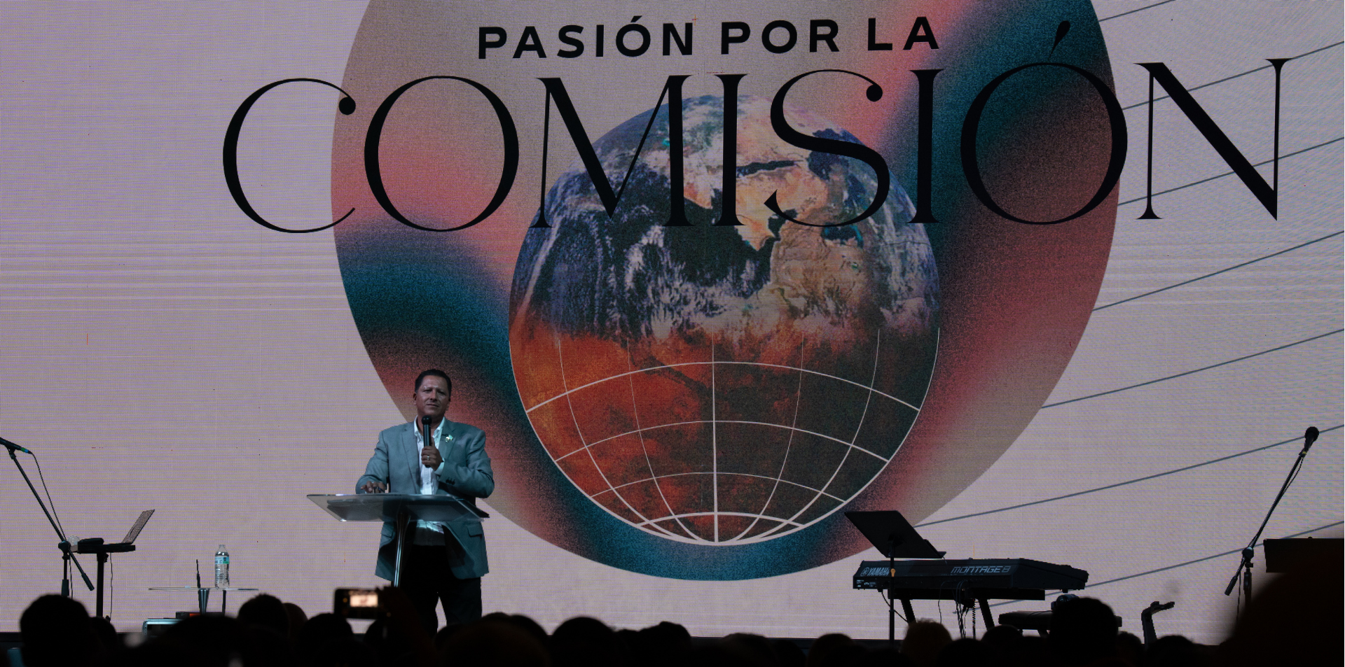 Hombre con traje azul dando un discurso en un escenario con proyección de la Tierra y el texto 'Pasión por la Comisión' y un fondo de plenario en un evento