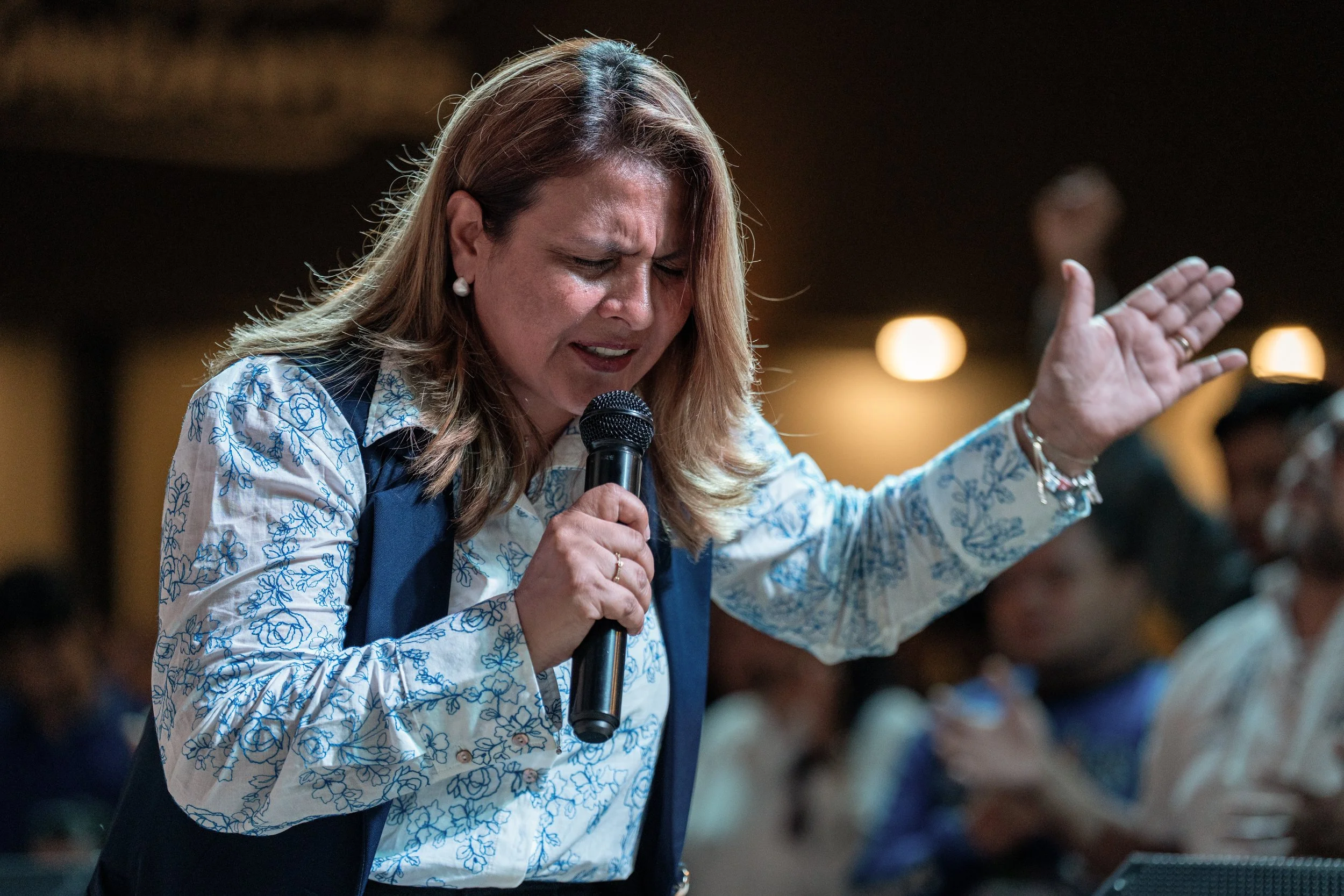 Mujer con blusa con diseño de flores sosteniendo un micrófono y con la vista cerrada en un discurso religioso o espiritual.