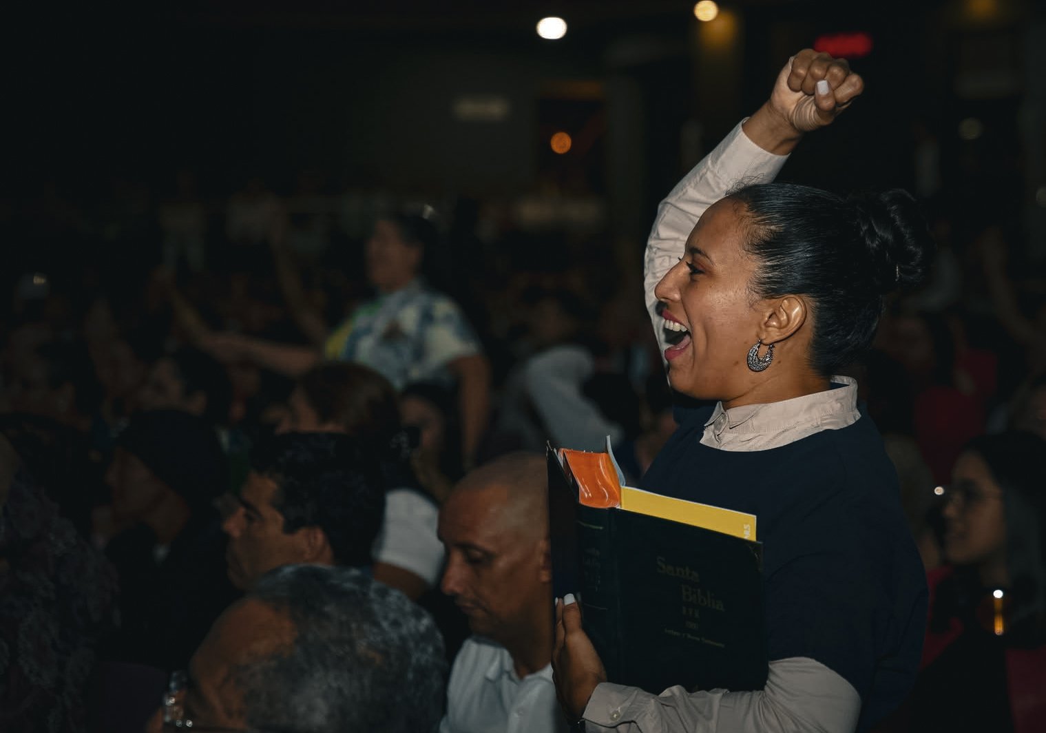 Mujer sonriendo con brazo en alto en una congregación, sosteniendo una Biblia y libros, en un ambiente oscuro con muchas personas