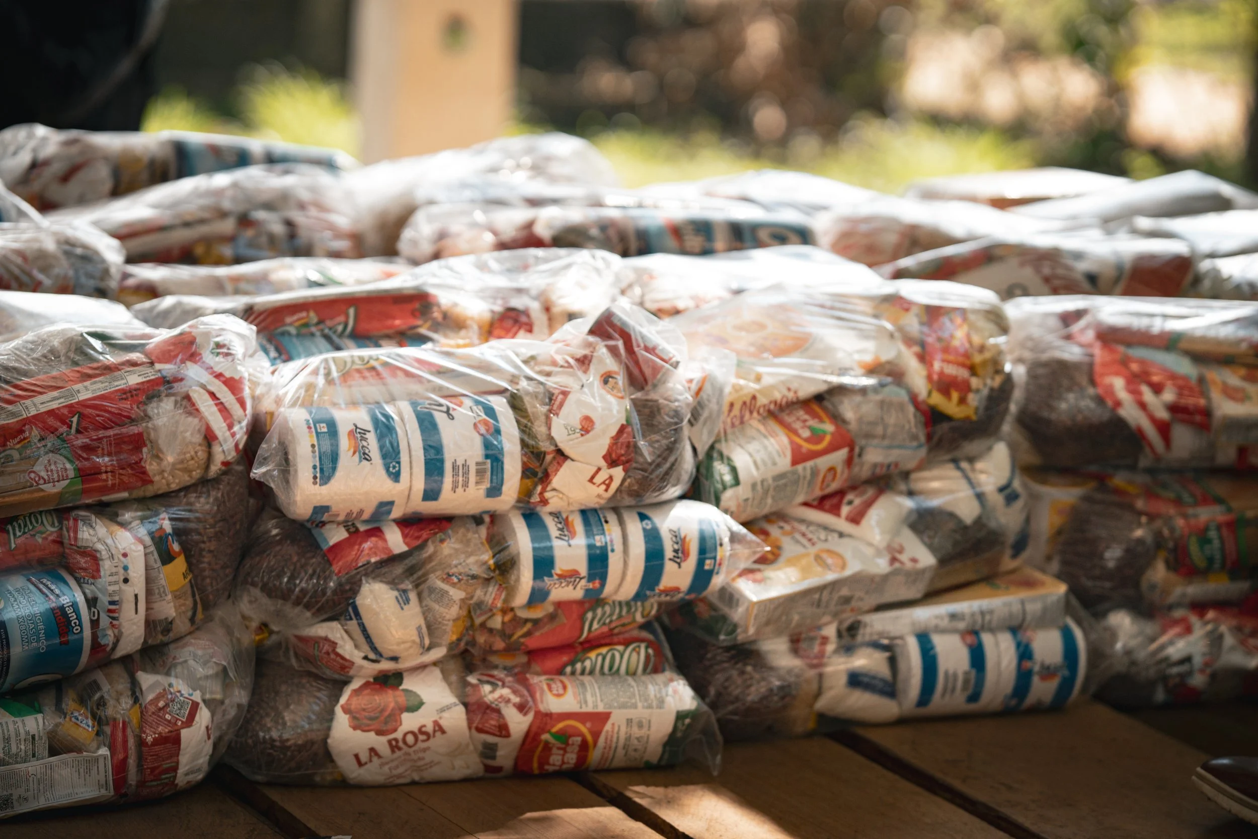 Montón de bolsas plásticas con productos alimenticios, en un entorno con luz natural y fondo difuso de árboles o plantas.