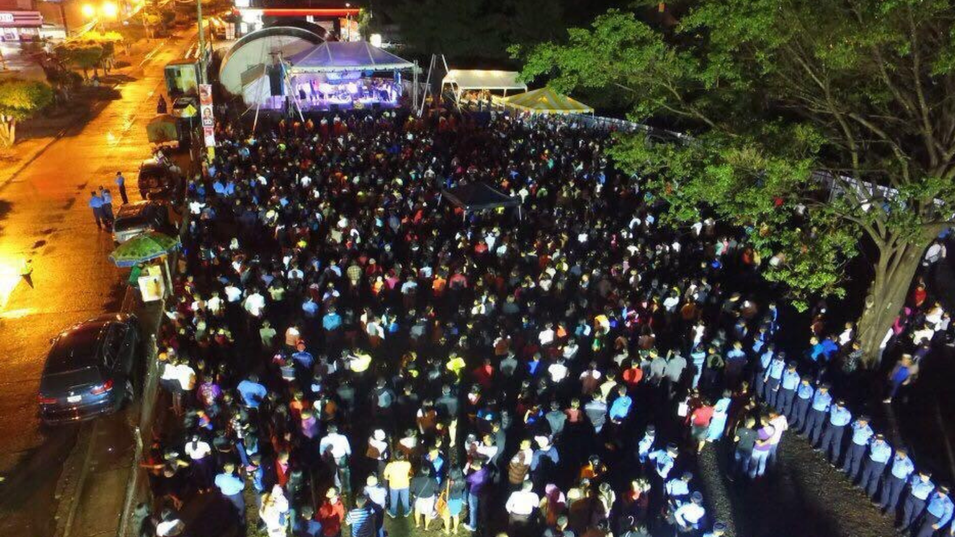 Gran multitud de personas asistiendo a un concierto al aire libre en la noche, con escenario iluminado y árboles a un lado.