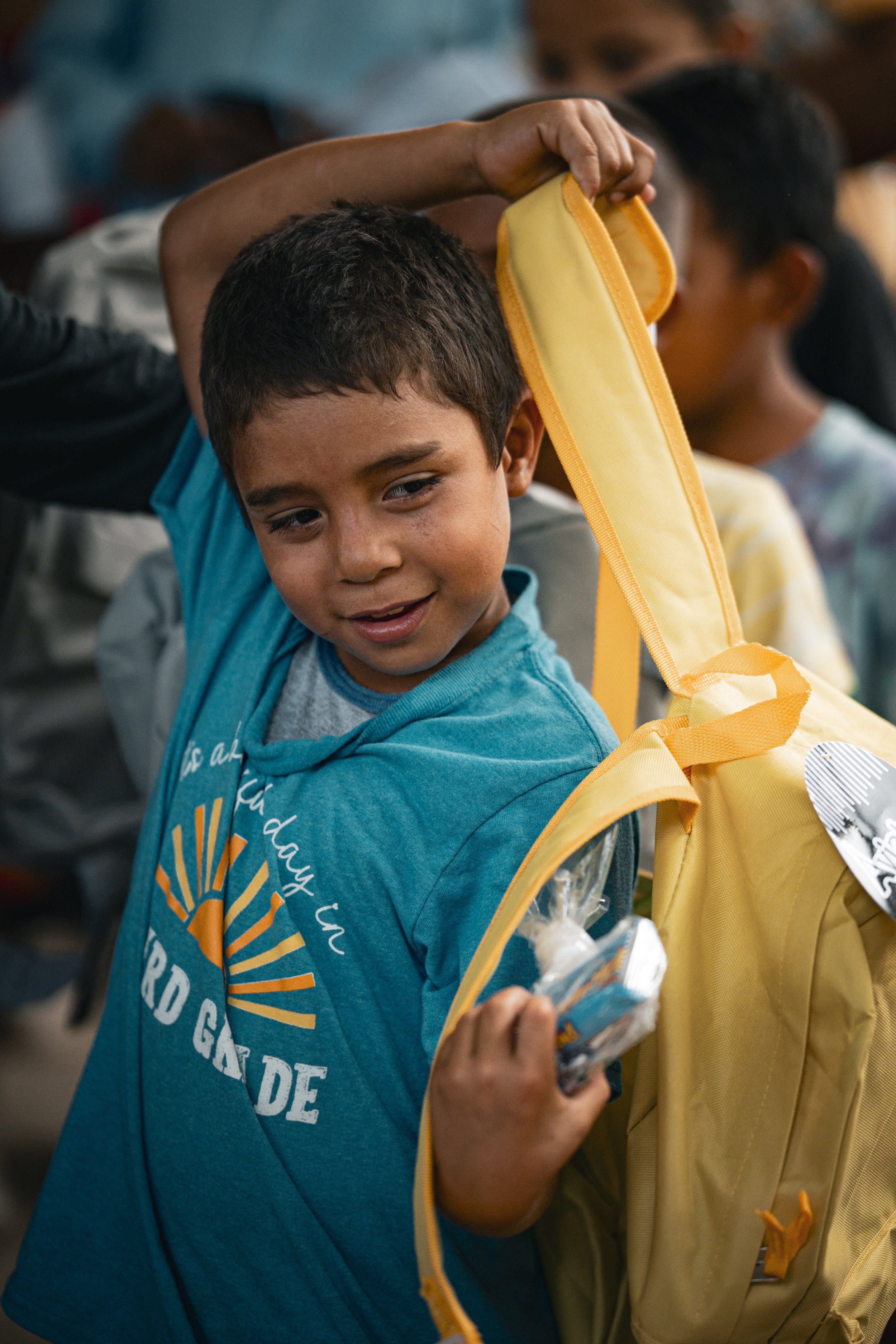 Un niño en una camiseta azul y mochila amarilla en un lugar concurrido, probablemente en un evento o escuela.