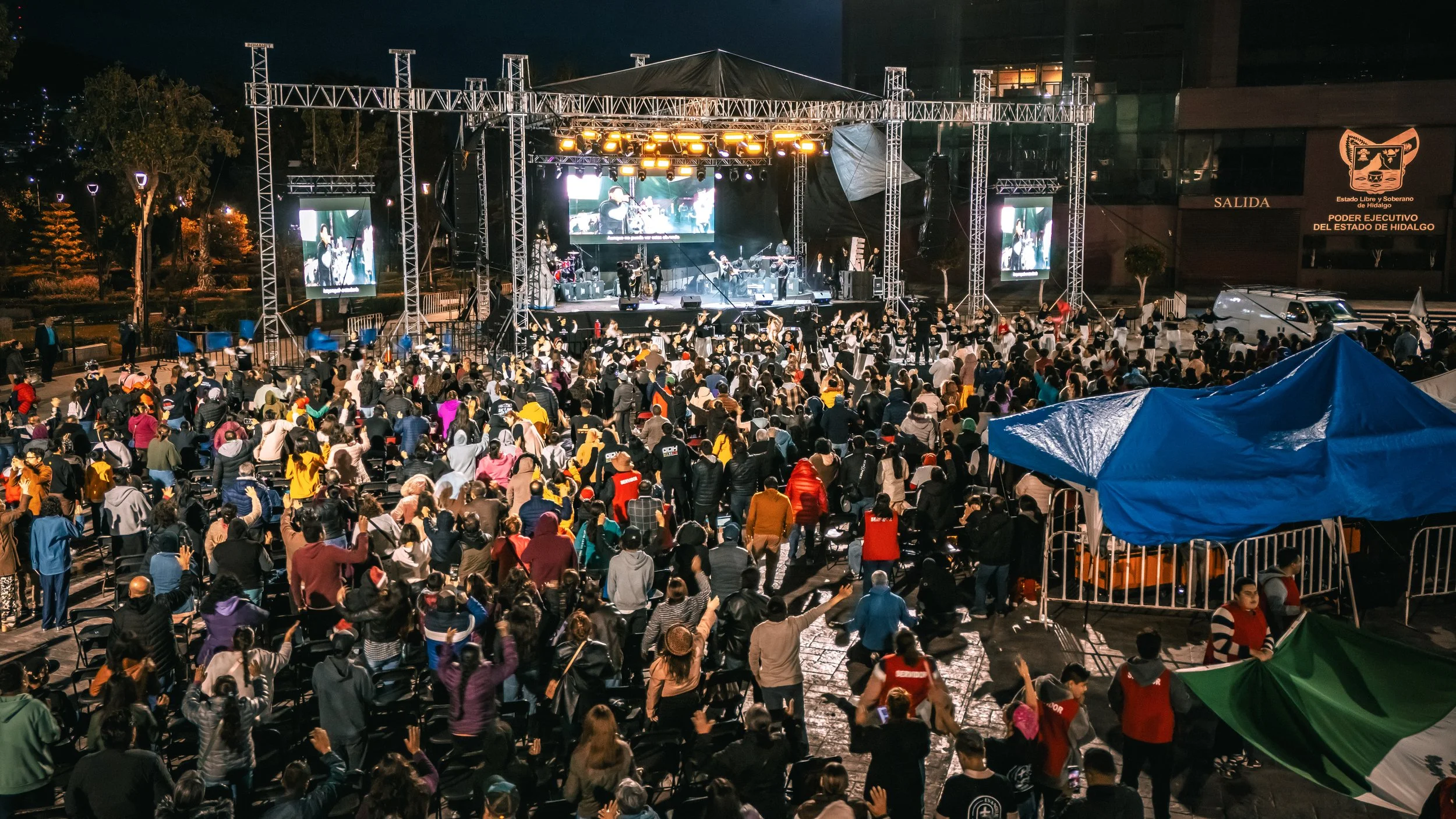 Concierto al aire libre en la noche con una multitud de personas disfrutando en un espacio abierto, escenario con músicos, pantallas grandes y estructura de iluminación.