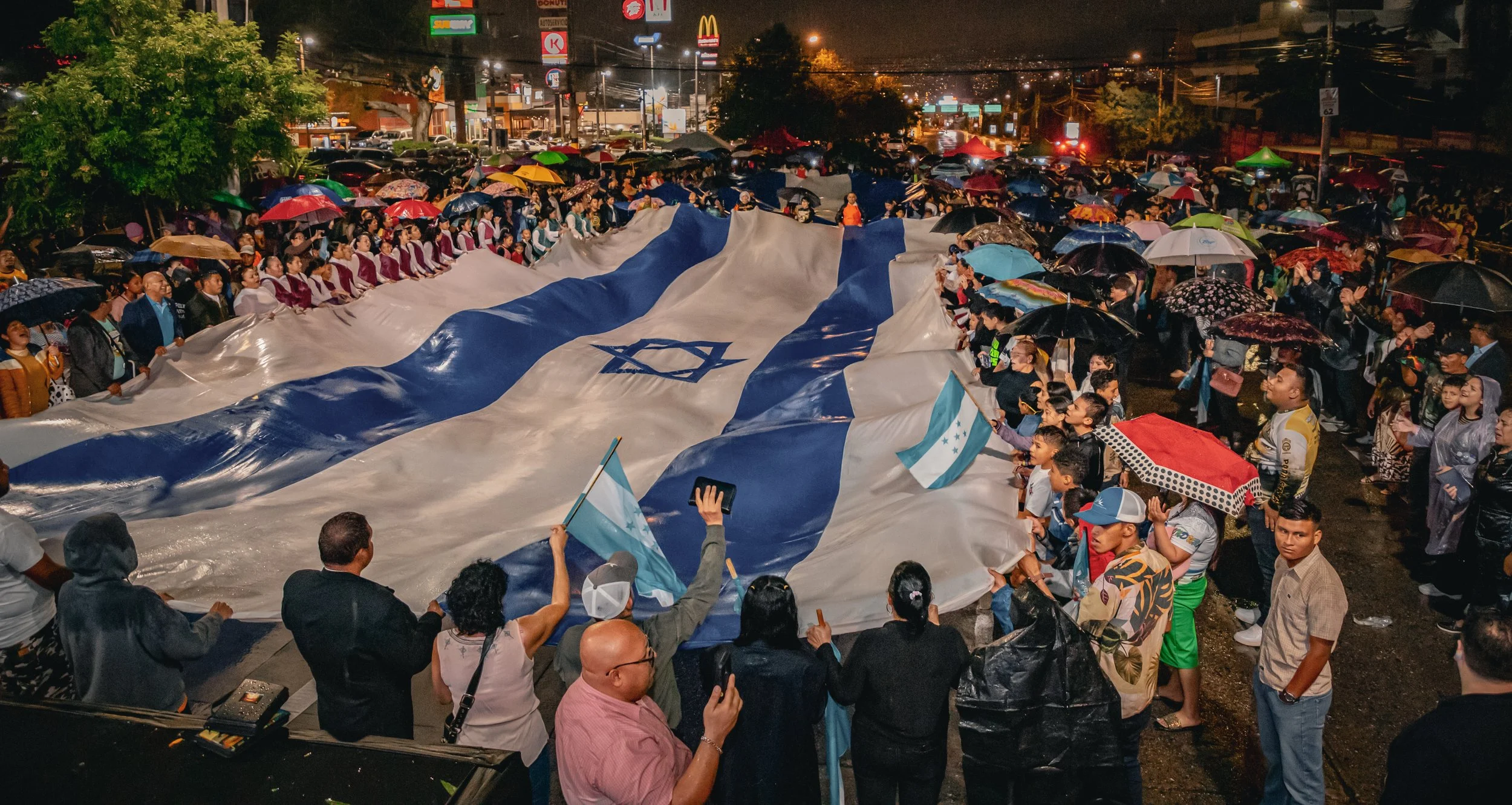 Grupo de personas en una marcha o protesta nocturna, portando una bandera grande de Honduras, con paraguas abiertos y rodeados de tiendas y signos luminosos en la ciudad.