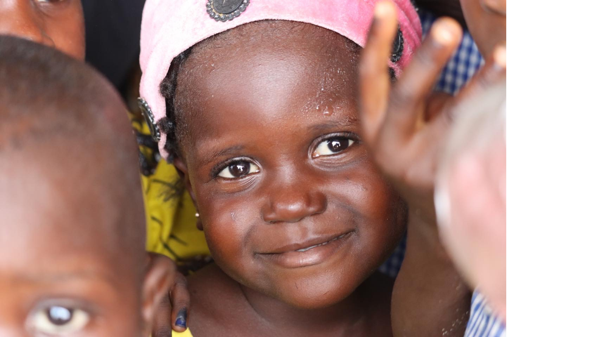 Niña africana con una sonrisa, rodeada de otras personas, con una gorra rosa, en un ambiente grupal.
