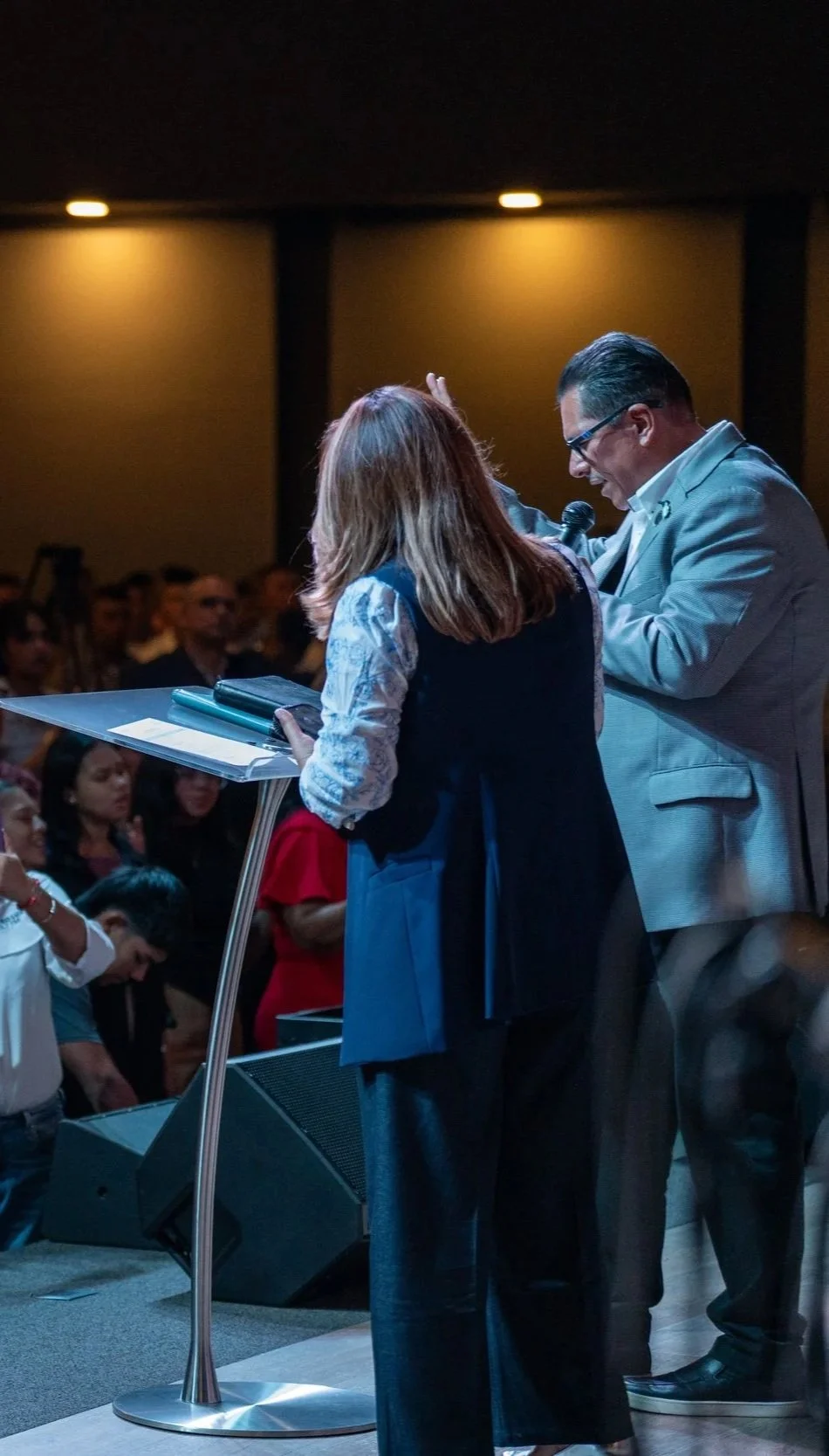 Dos personas en un escenario, una de ellas con micrófono, mientras una audiencia los observa.