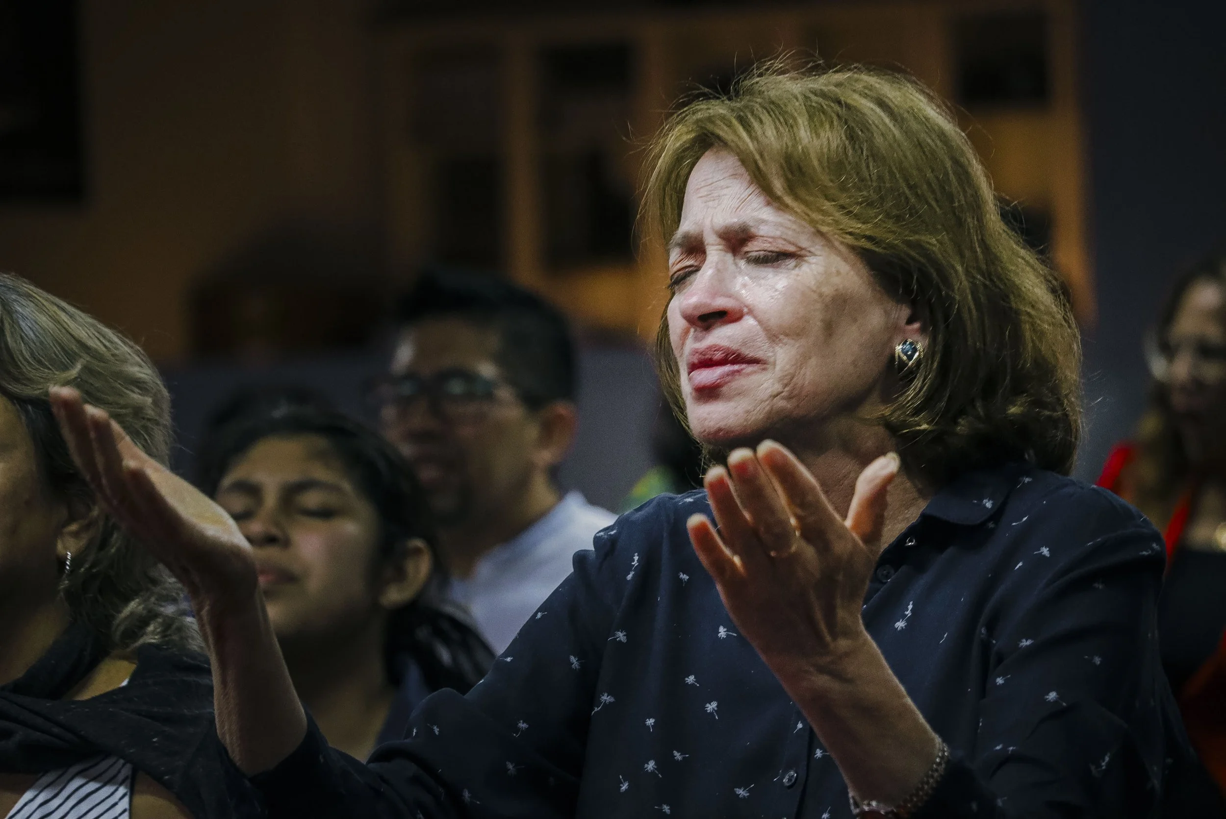 Mujer con cabello castaño claro y ojos cerrados en oración o meditación, rodeada de varias personas en un espacio interior, en un acto de plegaria o reflexión.