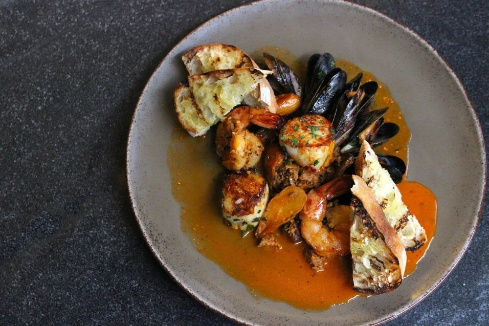 Seafood dish with shrimp, mussels, scallops, and toasted bread slices on a grey plate with sauce.